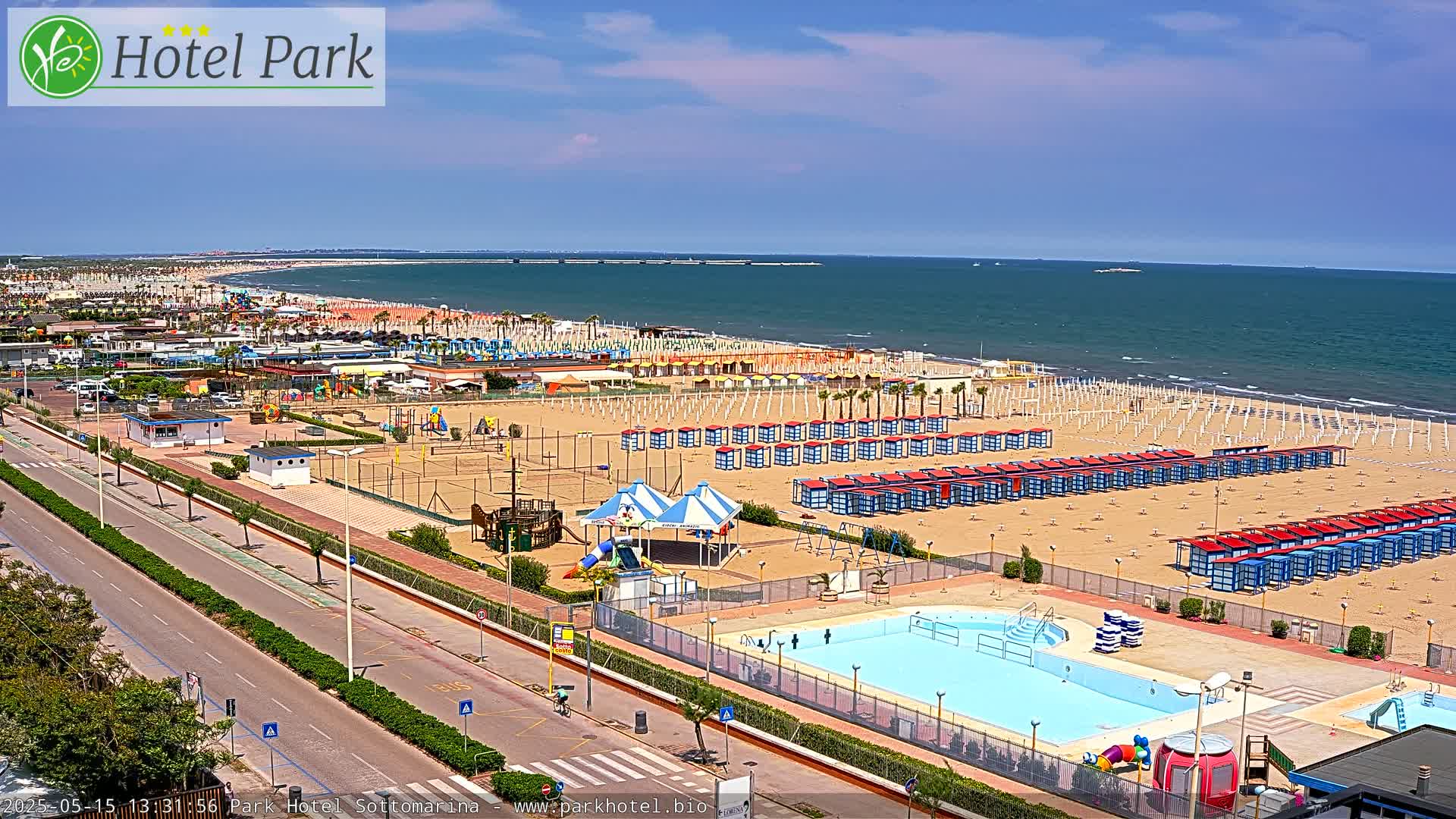 A sunny day at a beachside resort shows a long sandy beach with rows of beach huts, a swimming pool, playground equipment, and a road bordering the beach.