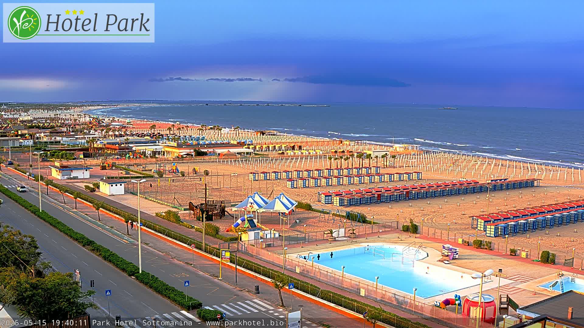 An aerial view of a beach with rows of beach cabanas, a swimming pool, playground equipment, and a road, under a partly cloudy sky.