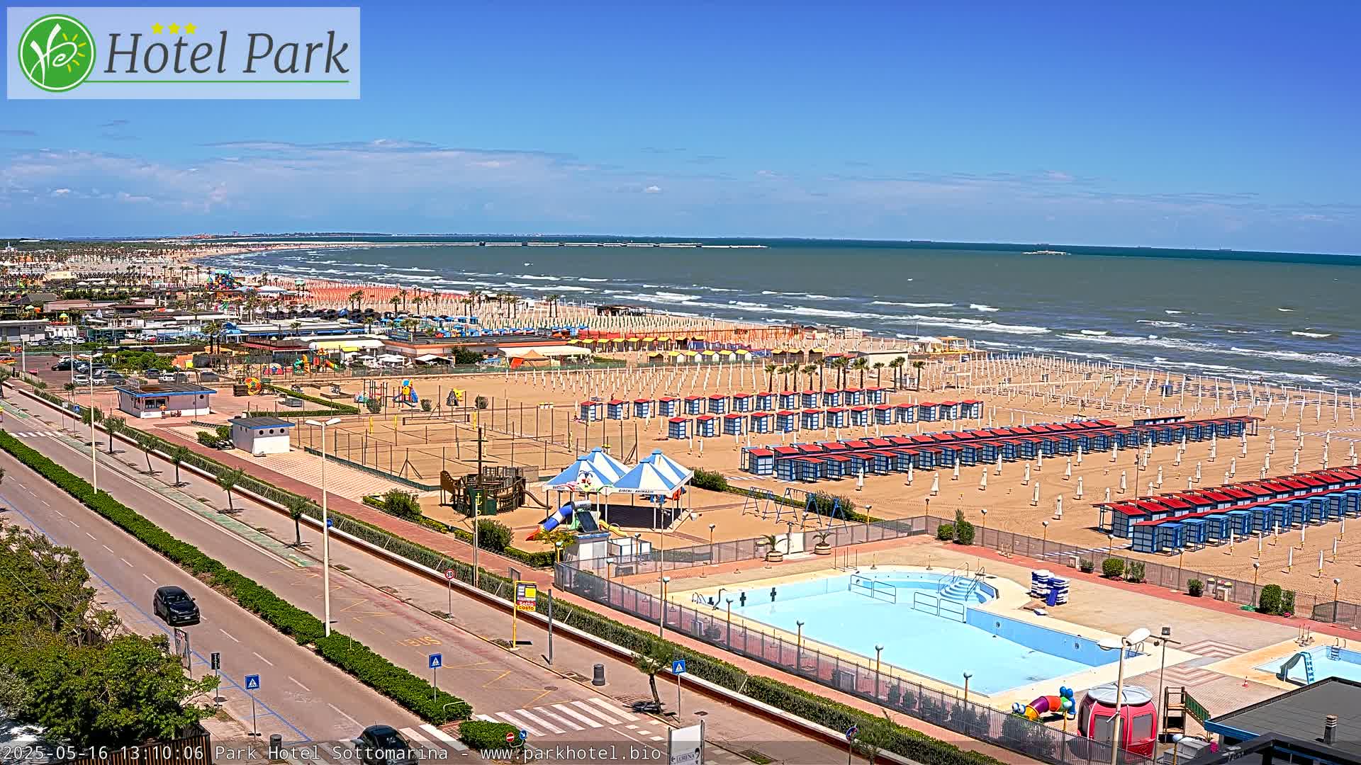 An aerial view shows a beach with numerous beach umbrellas and cabanas, a swimming pool, playground equipment, and a roadway, all under a sunny sky and with a moderately wavy ocean in the background.