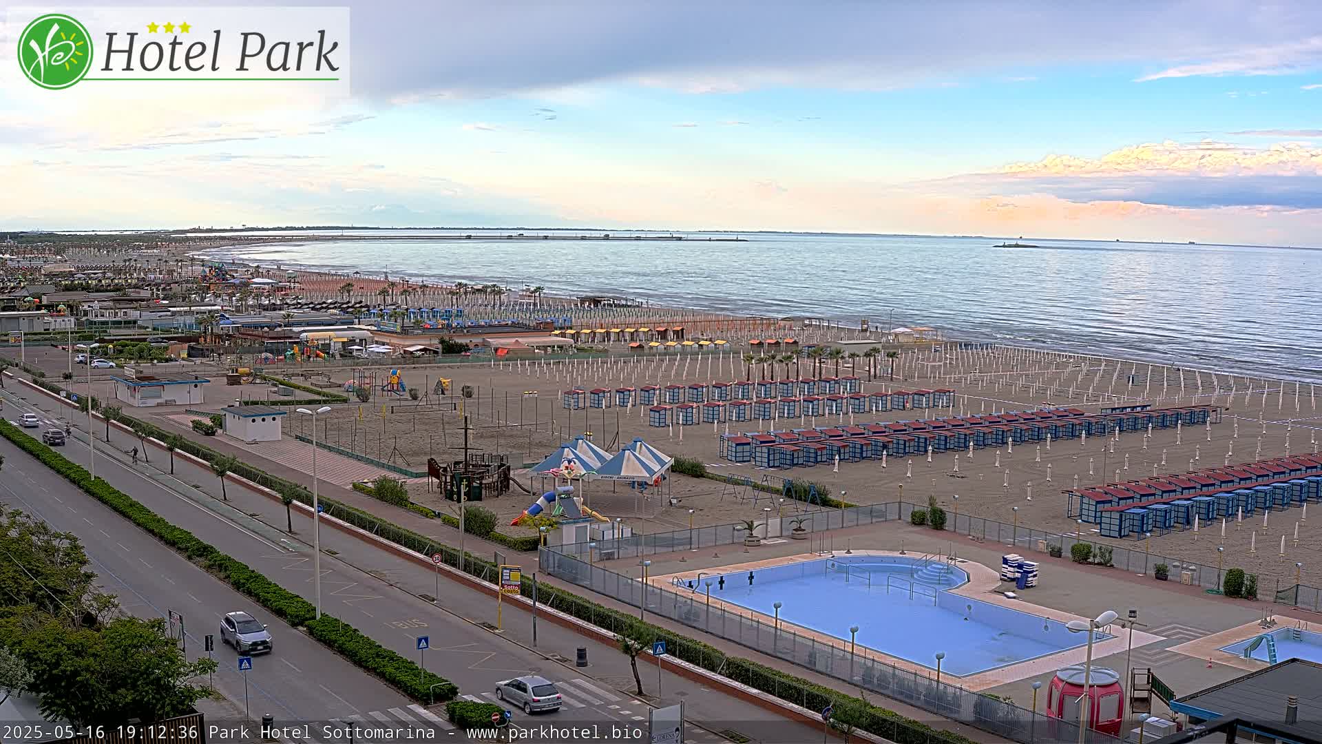 A sandy beach with numerous beach umbrellas and a swimming pool is seen next to a road, all under a partly cloudy sky.
