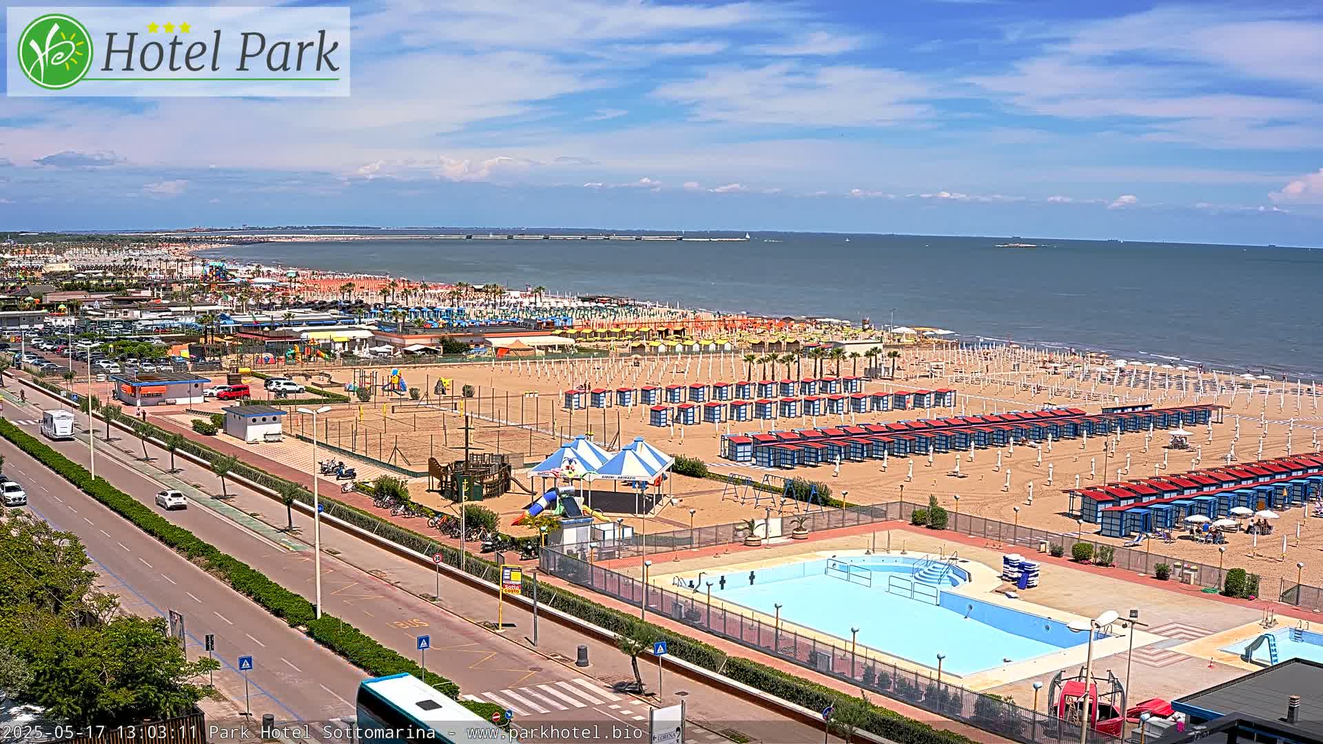 An aerial view of a beach scene on a sunny day shows a sandy beach with rows of beach umbrellas and changing rooms, a swimming pool, and a playground, all adjacent to a roadway and parking lot.