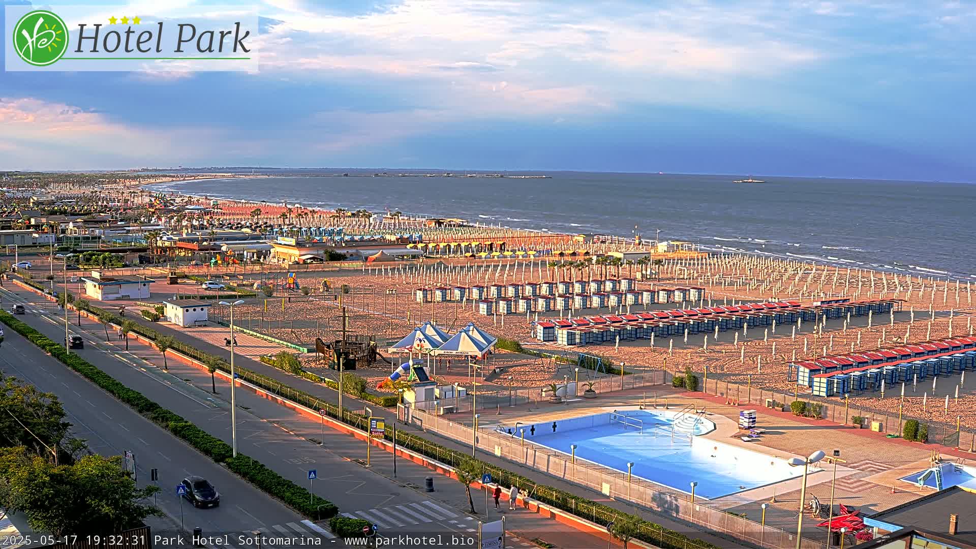 An aerial view shows a beach with rows of beach umbrellas and changing rooms, a swimming pool, and a road leading to the ocean under a partly cloudy sky.