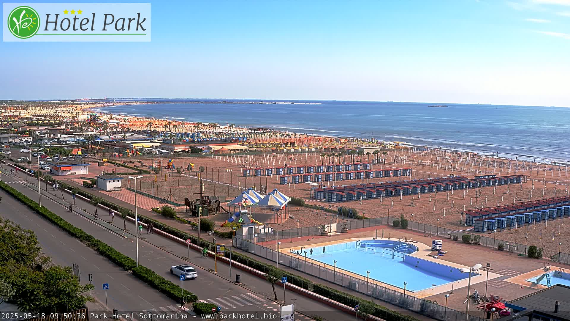 A beach with rows of beach cabanas, a swimming pool, playground, and parking lot is visible next to a road, all under a clear sunny sky and overlooking a calm ocean.