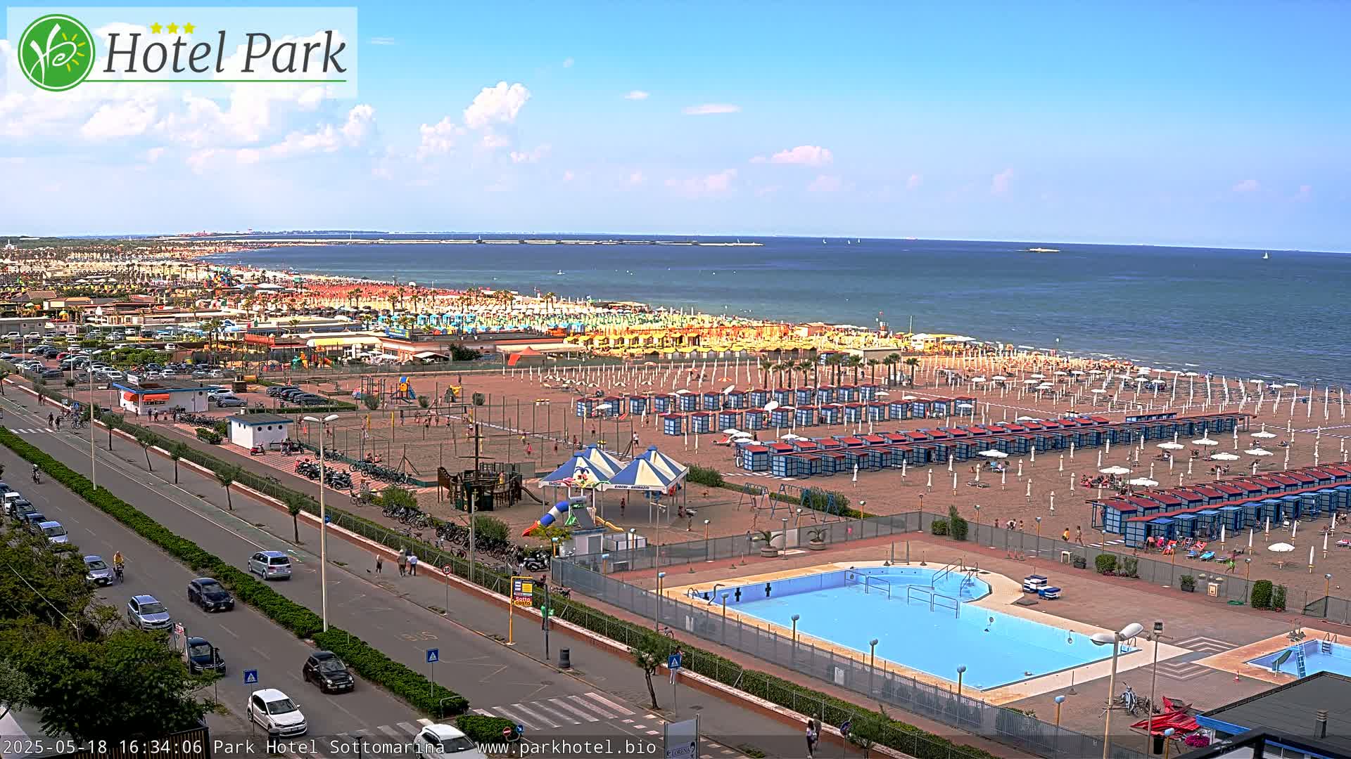 A beach scene on a partly sunny day shows a sandy beach with numerous beach umbrellas and cabanas, a swimming pool, and a roadway with cars.