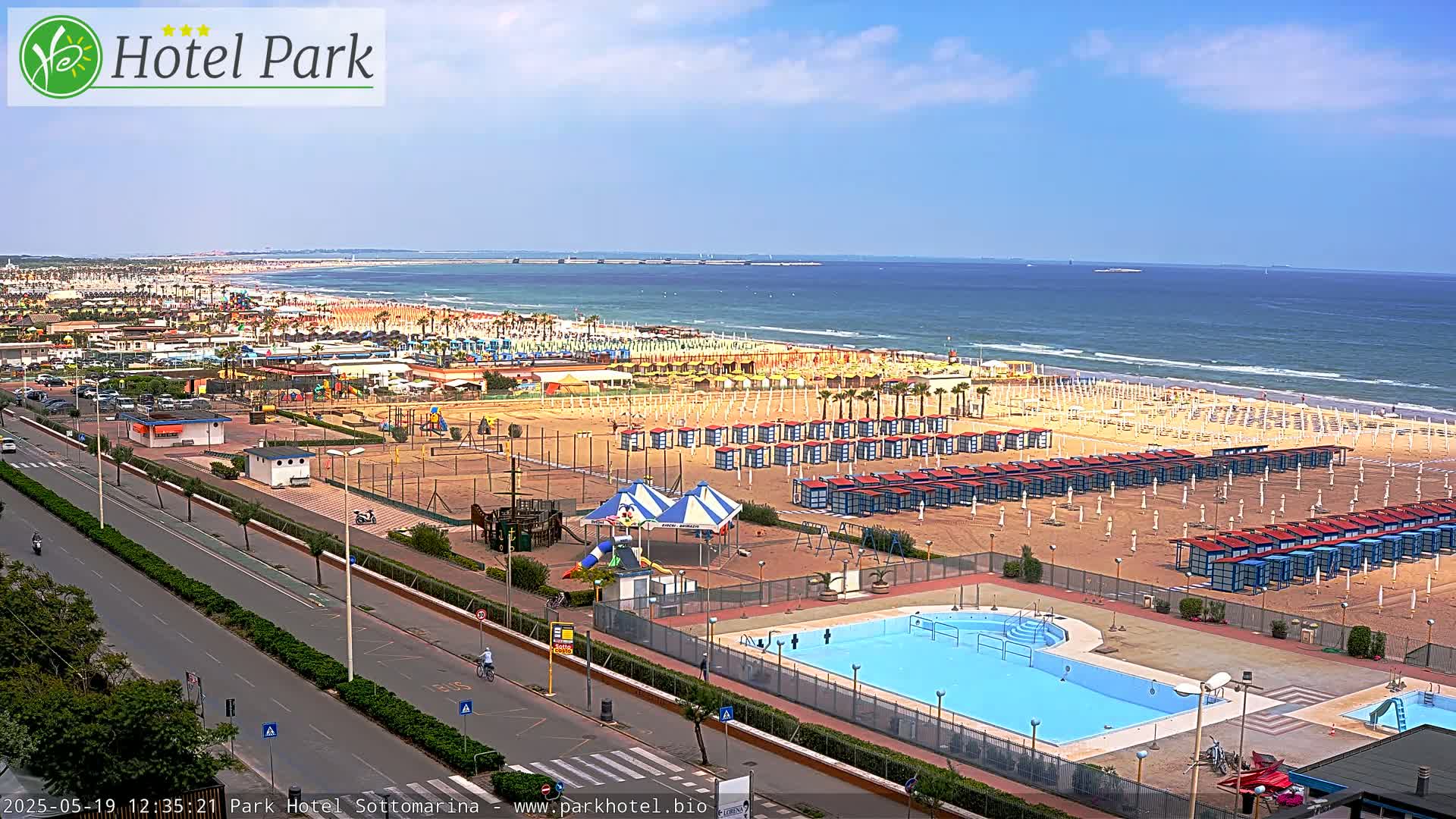 A beach with numerous beach umbrellas and a swimming pool is seen from an elevated vantage point on a sunny day, with a calm ocean visible in the background.