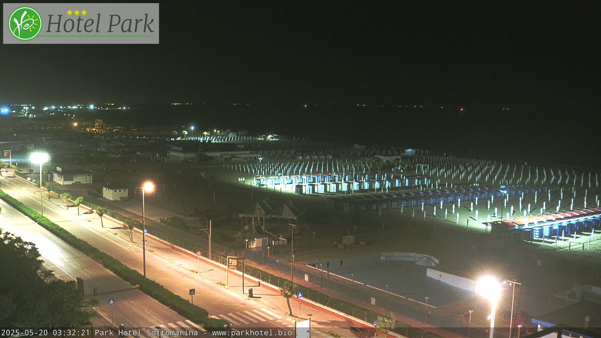 A nighttime, aerial view of a beach with rows of illuminated beach umbrellas and cabanas lining a dark ocean, adjacent to a brightly lit road.
