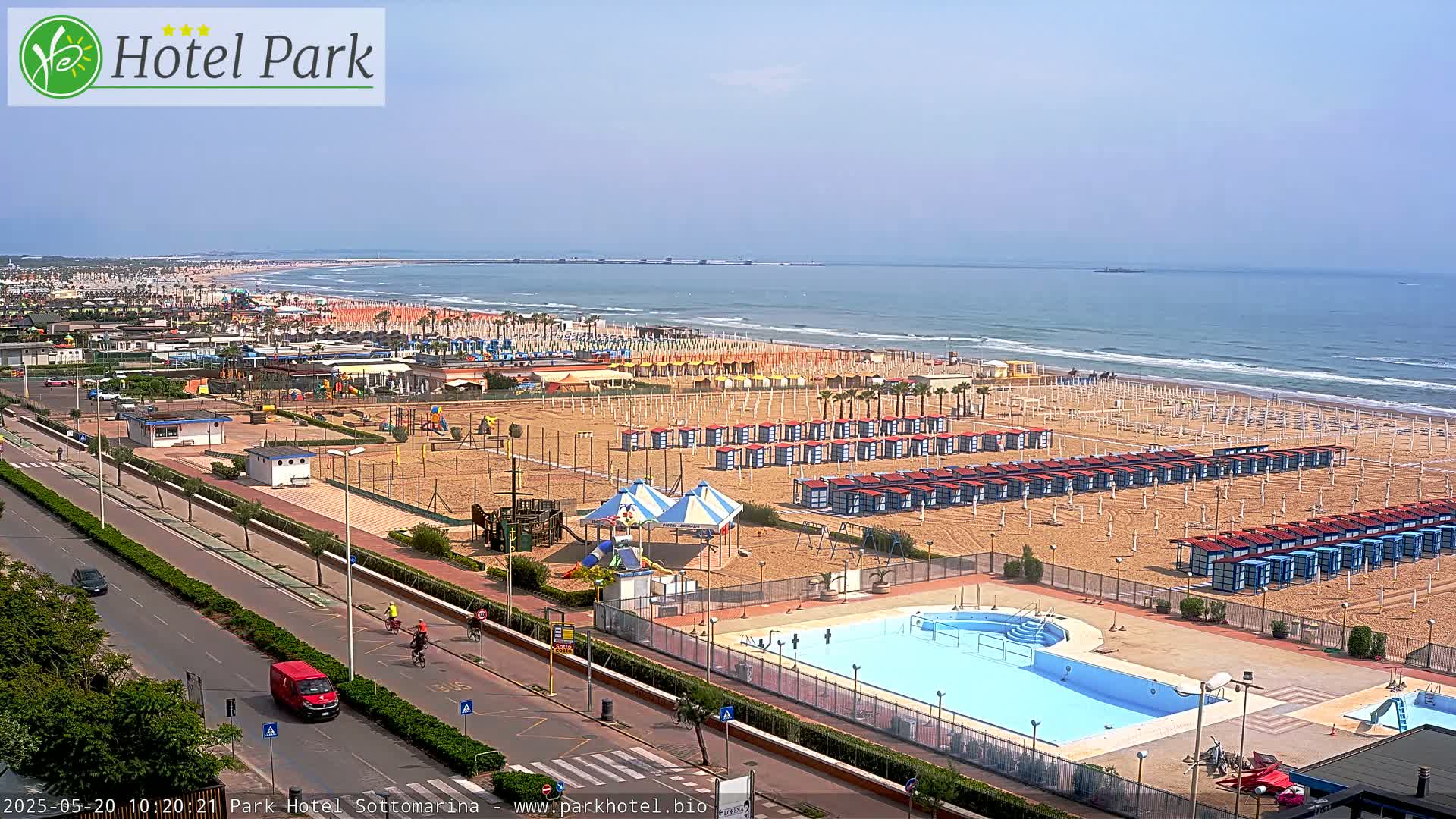 A sunny day at a beach resort shows a sandy beach with rows of beach huts, a swimming pool, playground equipment, and a road with some vehicles.