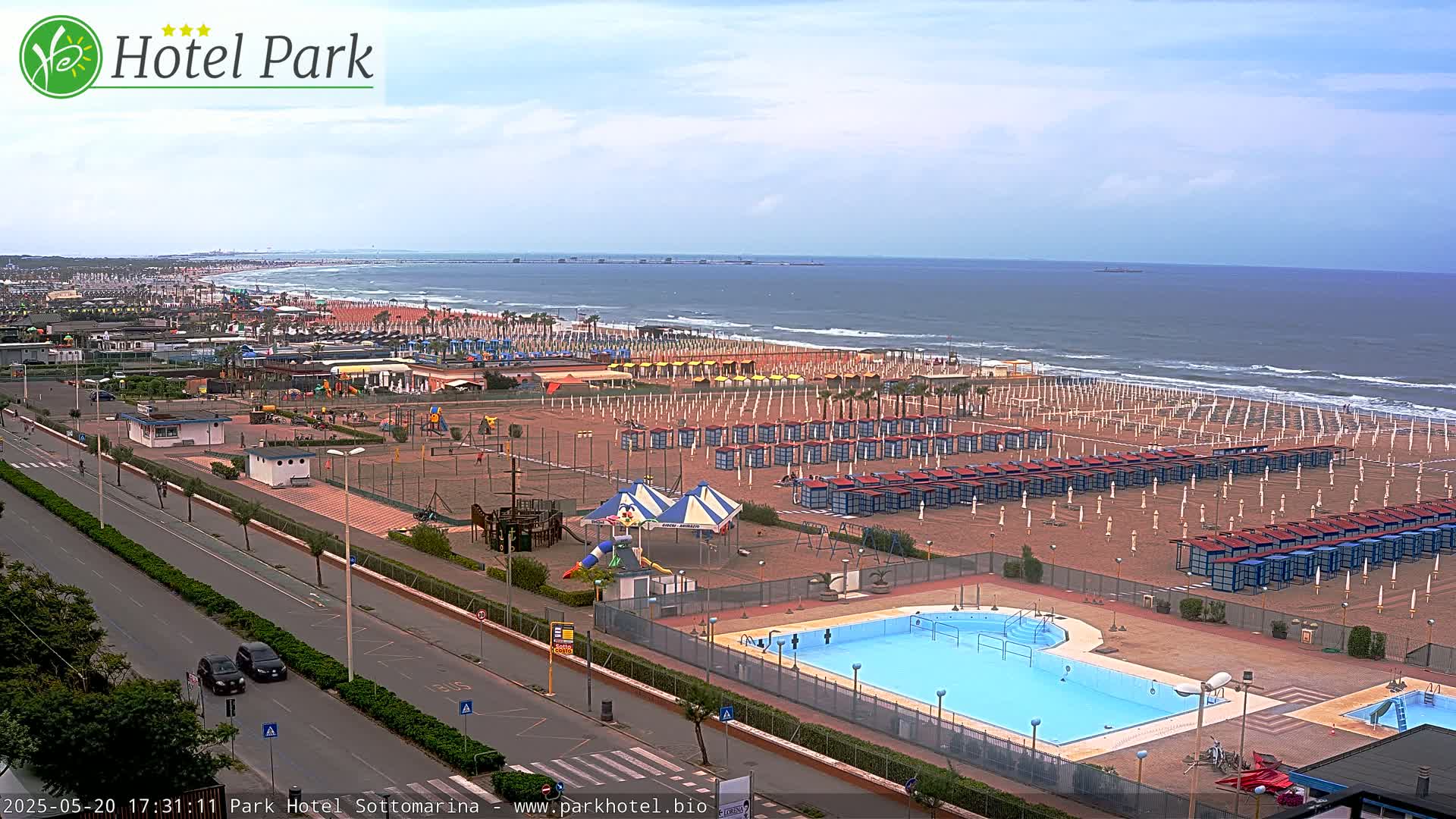An aerial view of a beach with numerous beach umbrellas and a swimming pool, under an overcast sky.