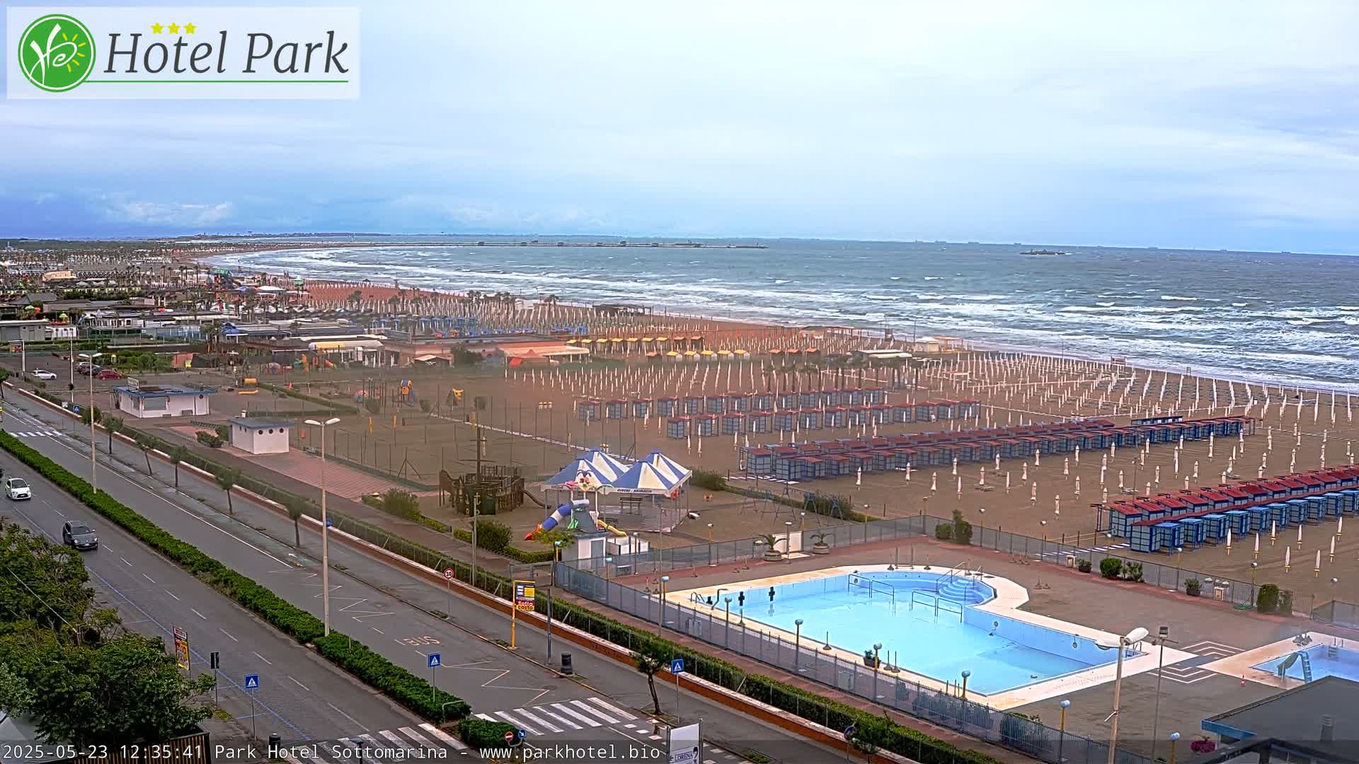 An aerial view shows a beach with rows of closed beach umbrellas and a swimming pool next to a road, all under a cloudy sky with a wavy ocean in the background.