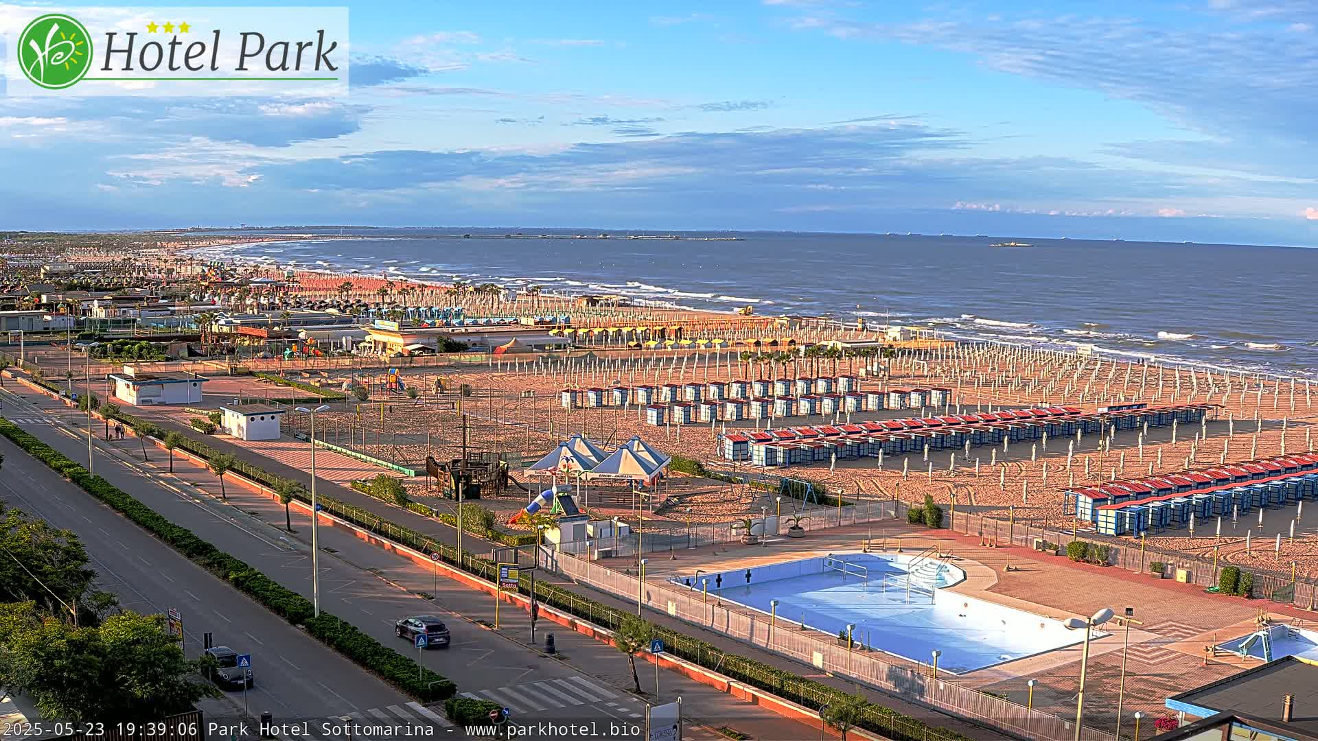 An expansive beach scene with numerous beach umbrellas and a swimming pool, under a partly cloudy sky.