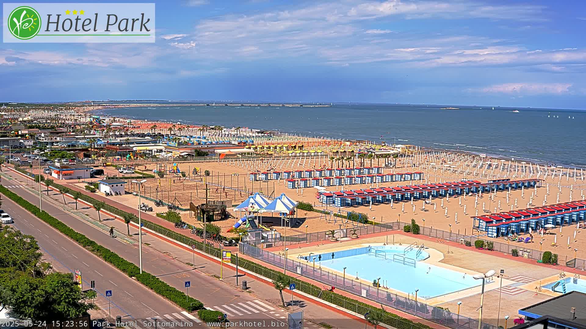 An aerial view shows a beach with numerous beach umbrellas and cabanas, a swimming pool, playground equipment, and a road, all under a mostly sunny sky.