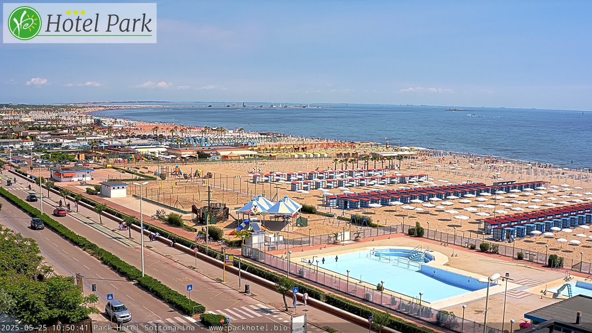 An aerial view of a beach with numerous umbrellas and beach huts, a swimming pool, playground, and a road, all under sunny skies.