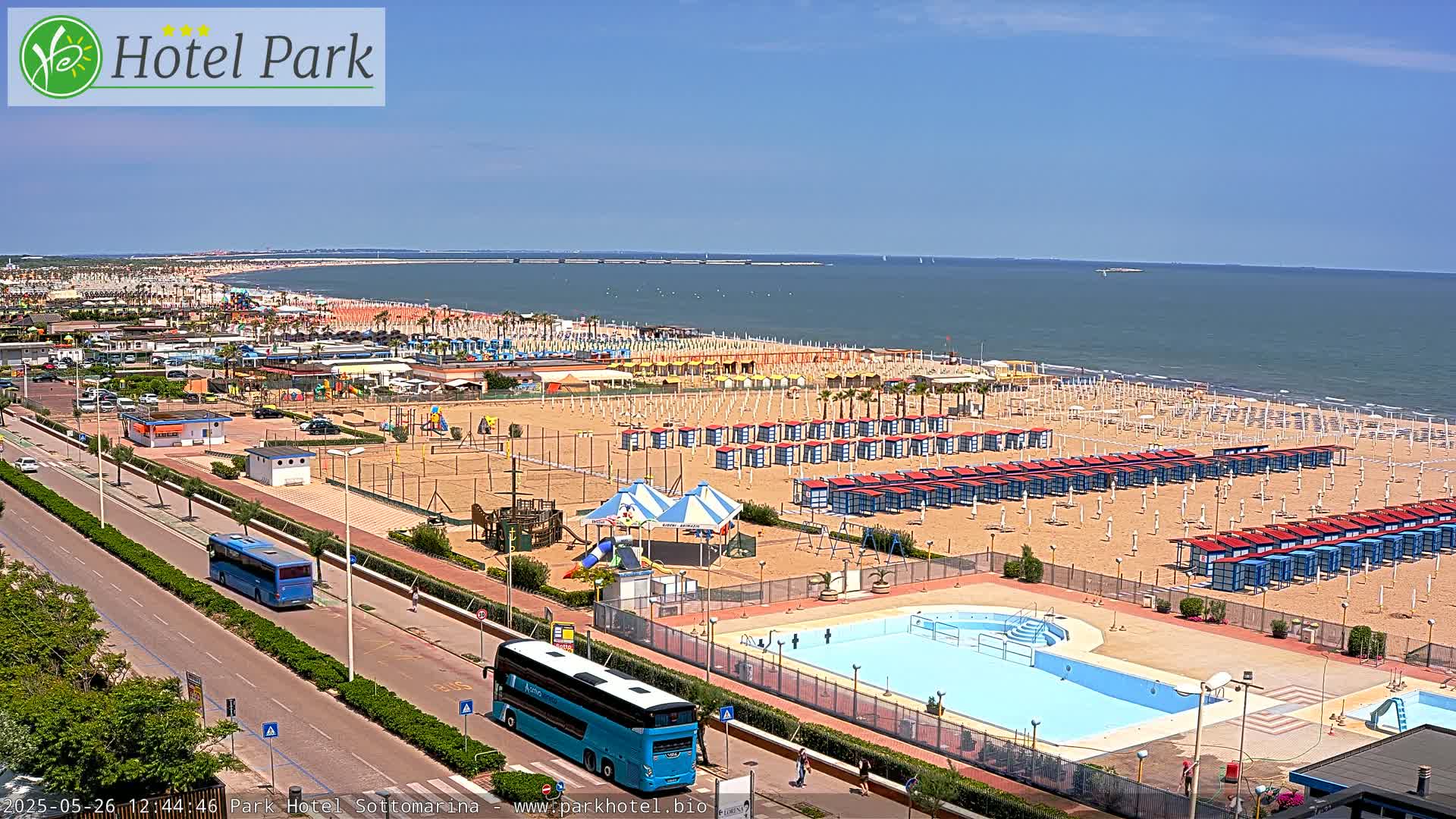 A sunny beachfront scene shows numerous beach umbrellas and cabanas, a swimming pool, and two buses parked on a road next to the beach.