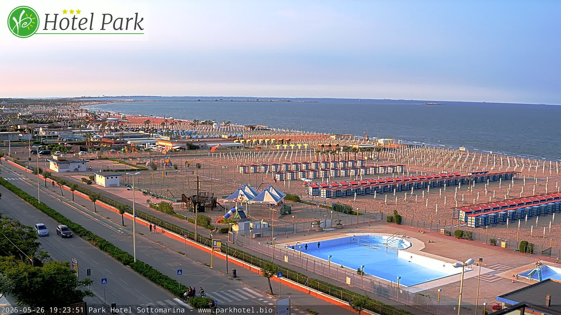 An aerial view shows a beach with rows of beach umbrellas and cabanas, a swimming pool, playground equipment, and a road next to the ocean under a clear, light-blue sky.