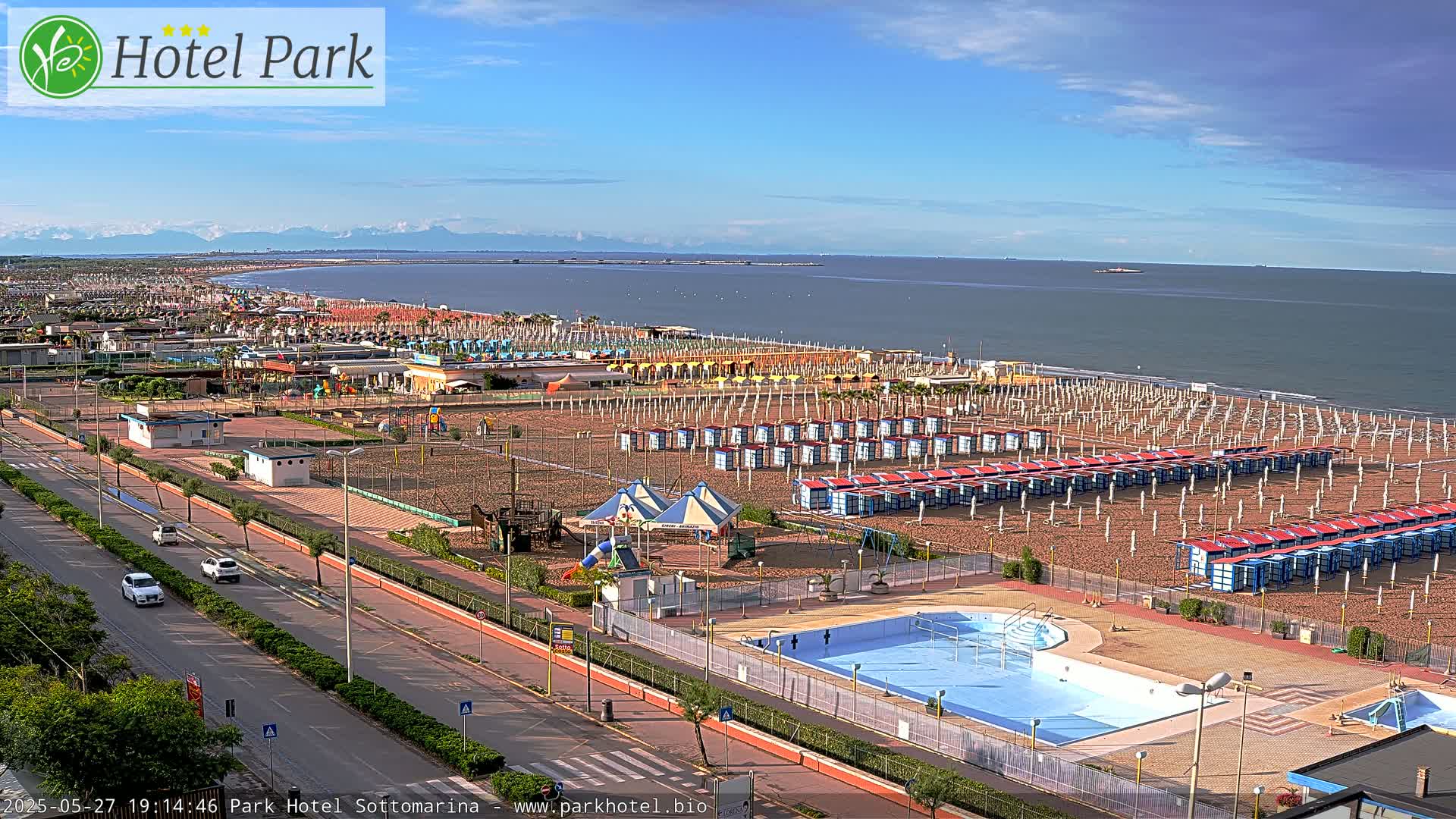 A beach with numerous beach umbrellas and changing rooms, a swimming pool, and a playground, overlooks a calm sea under a partly cloudy sky with mountains visible in the distance.