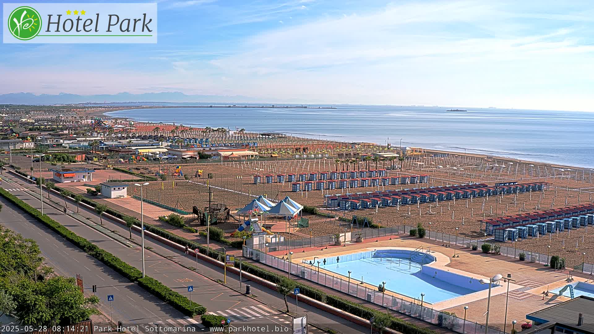 An aerial view of a beach resort on a sunny day shows a swimming pool, beach cabanas, playground equipment, and a road leading to the ocean.