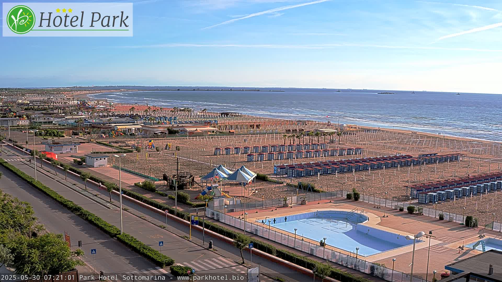 A beach with numerous beach huts, a swimming pool, and a playground is seen from an elevated vantage point on a sunny day with a calm ocean in the background.
