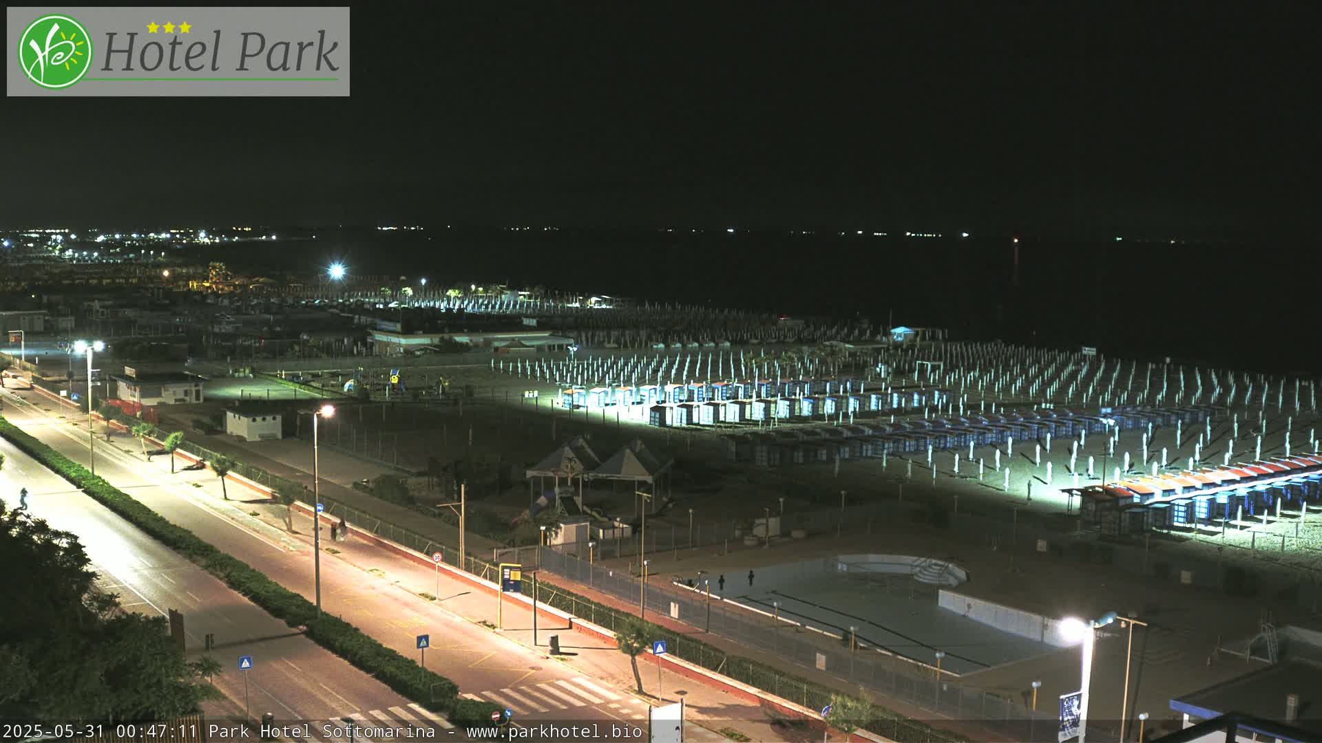 A nighttime view of a beach with rows of closed beach umbrellas and cabanas, illuminated along a roadway, next to a dark body of water.