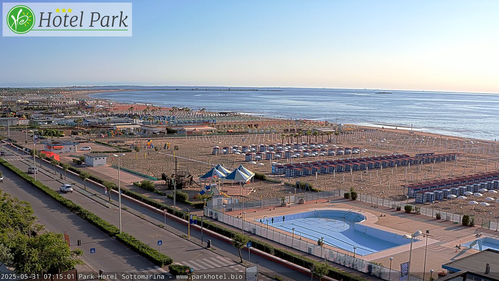 An aerial view shows a beach with numerous beach umbrellas and lounge chairs, a swimming pool, and a road with cars, all under a clear, sunny sky.