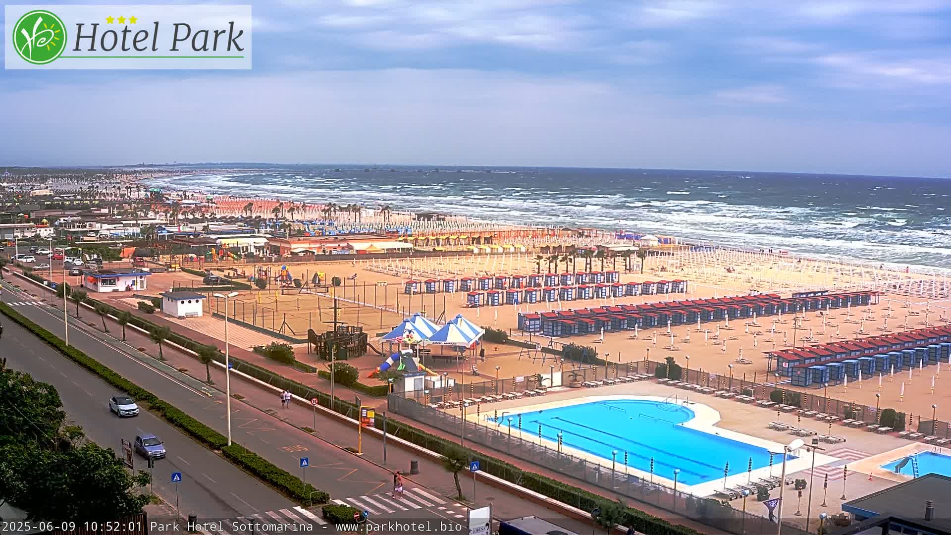 A beach with numerous beach umbrellas and a swimming pool is visible, next to a road with cars, all under a somewhat cloudy sky with a windy, wavy ocean in the background.
