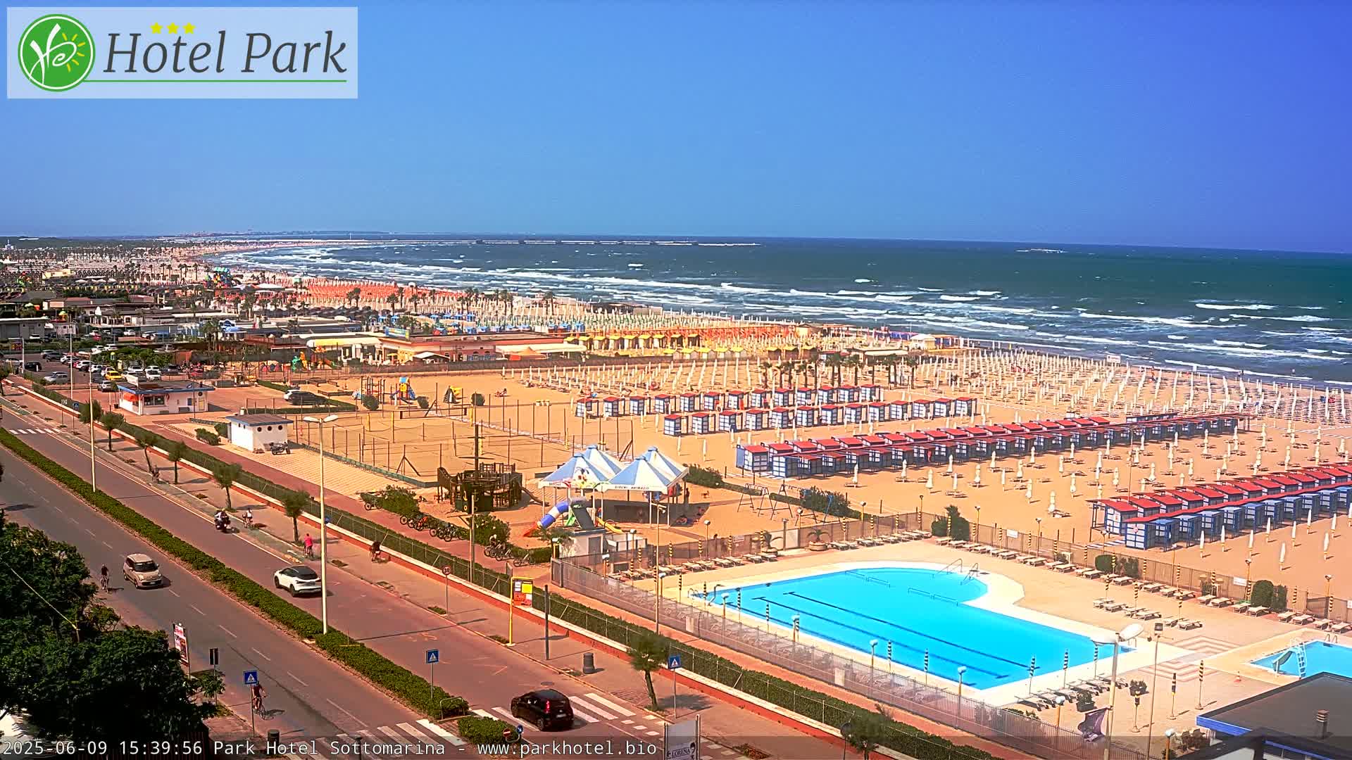 An aerial view shows a beach with numerous umbrellas and beach huts, a swimming pool, and a road with cars, all under a clear blue sky.