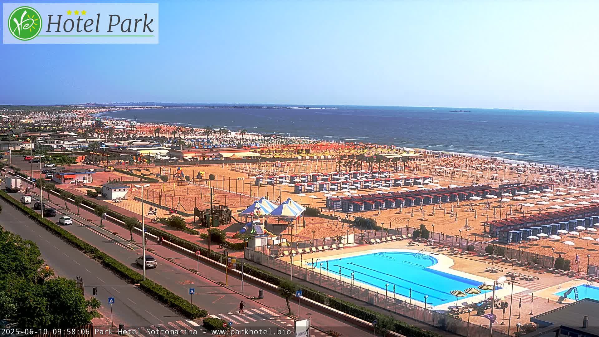 An aerial view shows a sunny beach with numerous umbrellas and beach chairs, a swimming pool, and a playground area next to a road with cars.