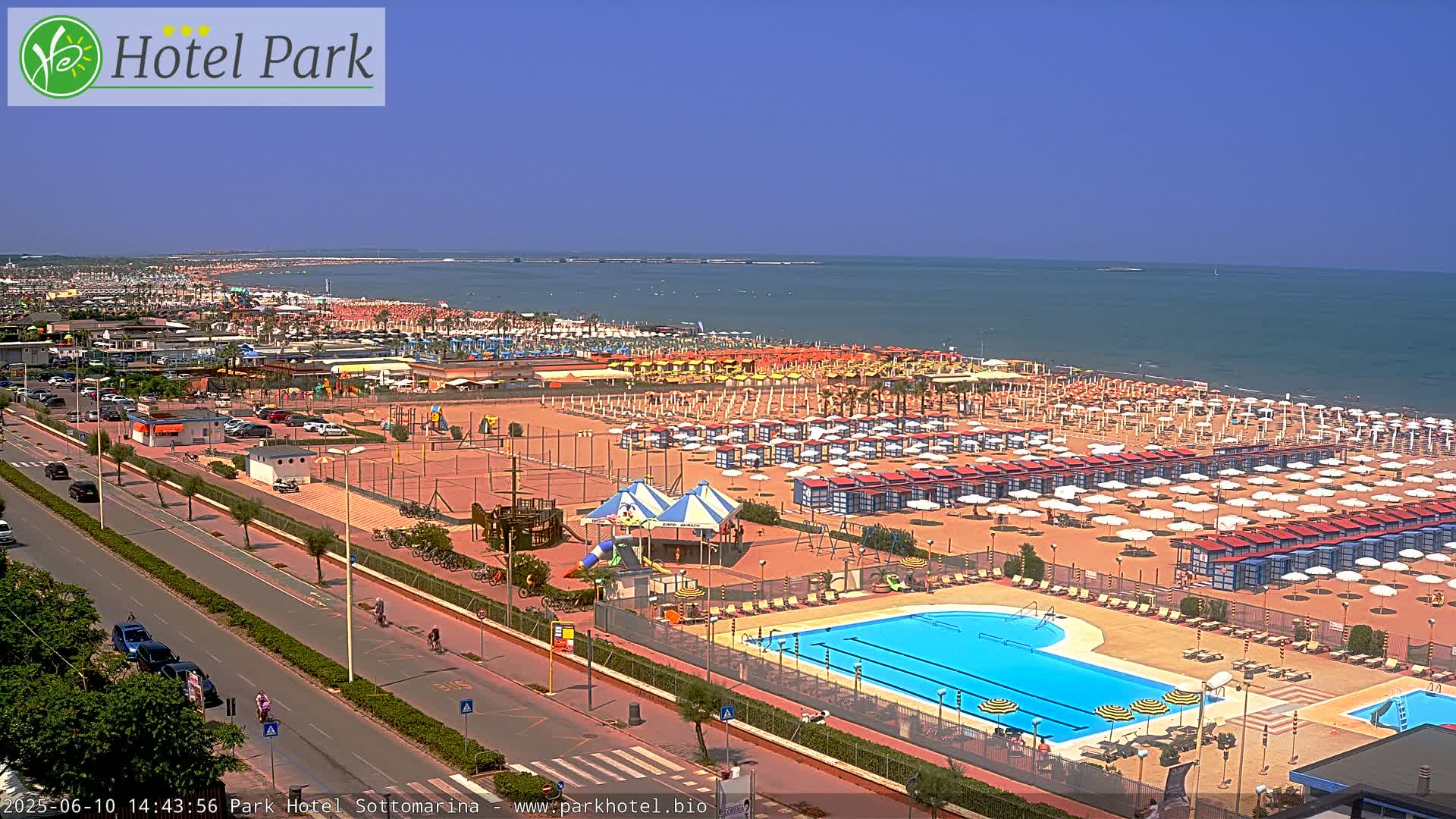 A sunny day at a beach resort showing a long sandy beach lined with umbrellas and beach chairs, a swimming pool, and a playground, all next to a road.