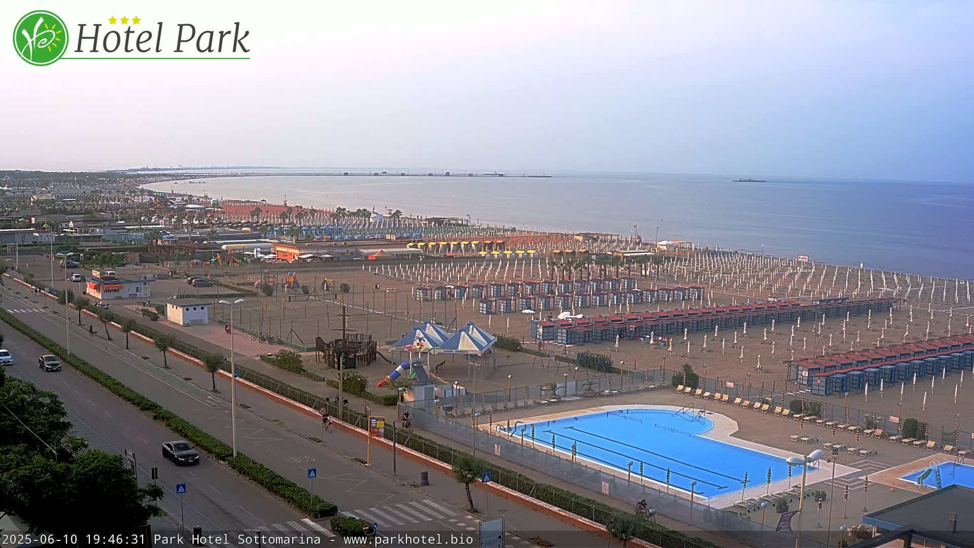 An aerial view of a beach resort shows a sandy beach with rows of beach umbrellas and chairs, a swimming pool, a playground, and a road leading to the ocean under a hazy sky.