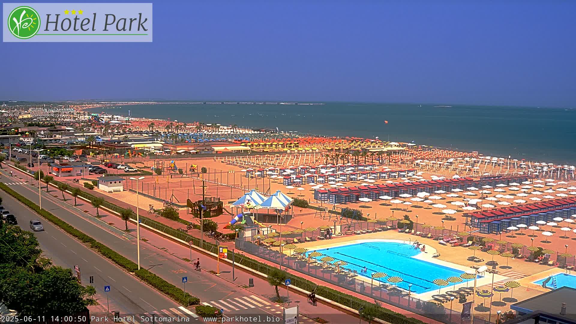 A sunny aerial view shows a beach with numerous umbrellas and beach chairs, a swimming pool, and a playground area adjacent to a road.