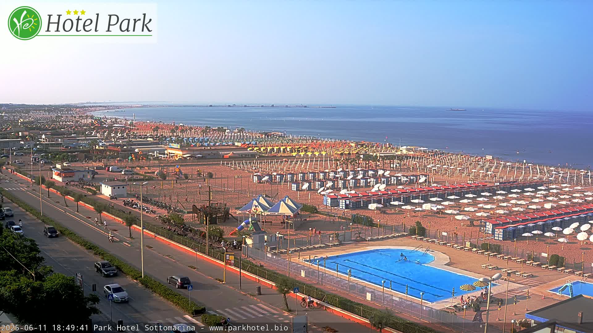 An aerial view of a beach with numerous umbrellas and beach cabanas, a swimming pool, a playground, and a road with cars, all under a clear, sunny sky.