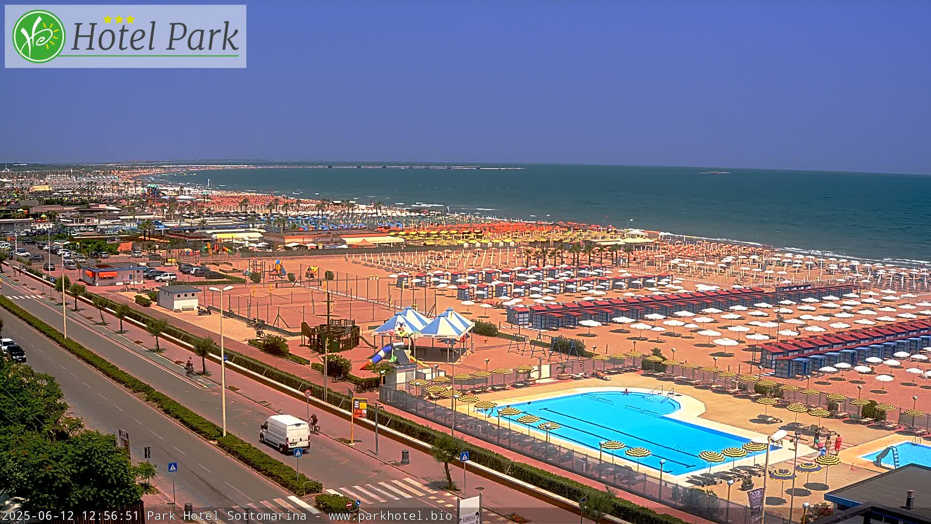 An aerial view of a beach resort on a sunny day, showing a sandy beach lined with umbrellas, a swimming pool, and a playground, all next to a road with some cars and a van.