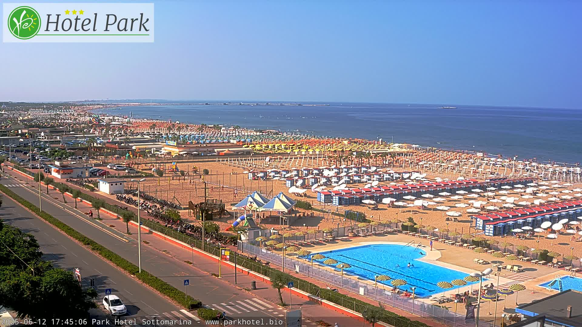 A sunny beach scene with numerous umbrellas and beach chairs, a swimming pool, and a road leading to the ocean.
