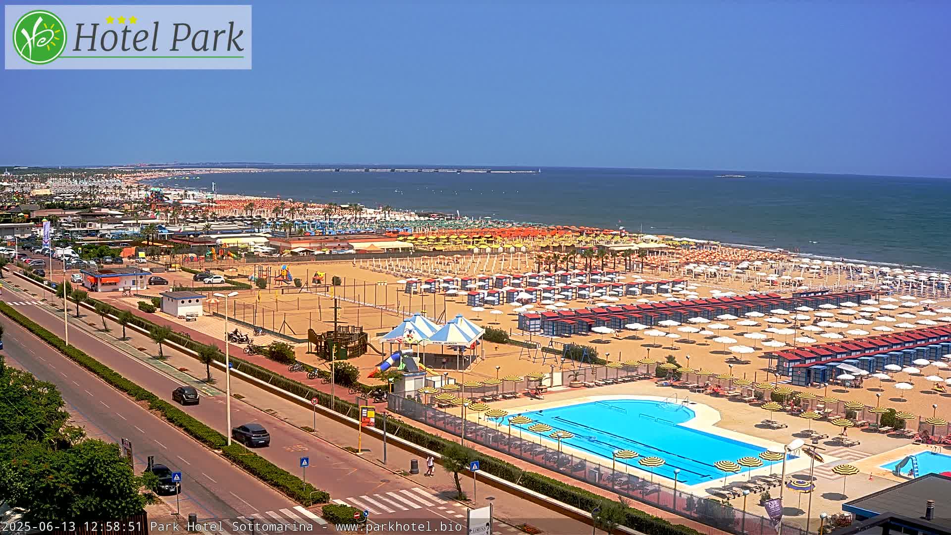 A sunny day at a beach resort shows a large sandy beach with numerous umbrellas and beach cabanas, a swimming pool, and playground equipment, all next to a road with cars.