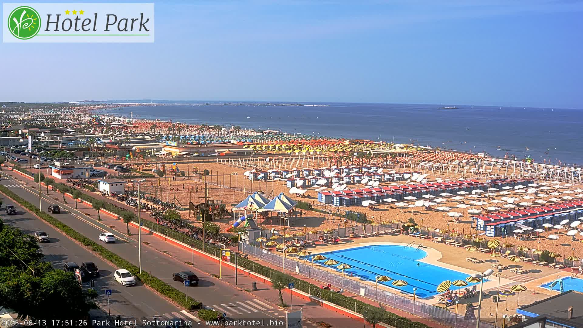 A sandy beach with numerous umbrellas and beach huts, a swimming pool, and a roadway is seen on a sunny day next to a calm ocean.