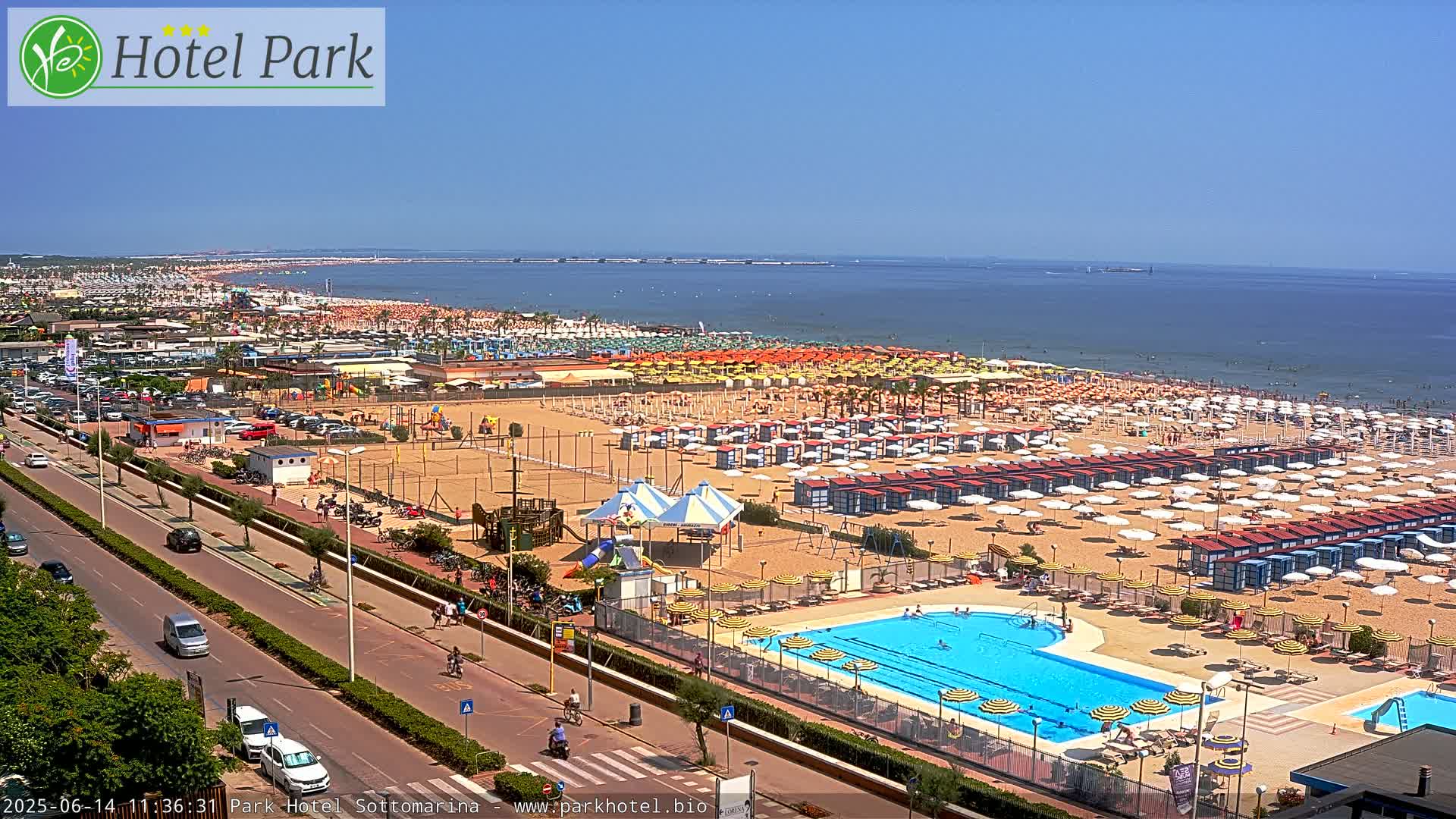 A sunny day at a beach resort shows a crowded sandy beach with numerous umbrellas and beach cabanas, a swimming pool, and a road lined with parked cars.