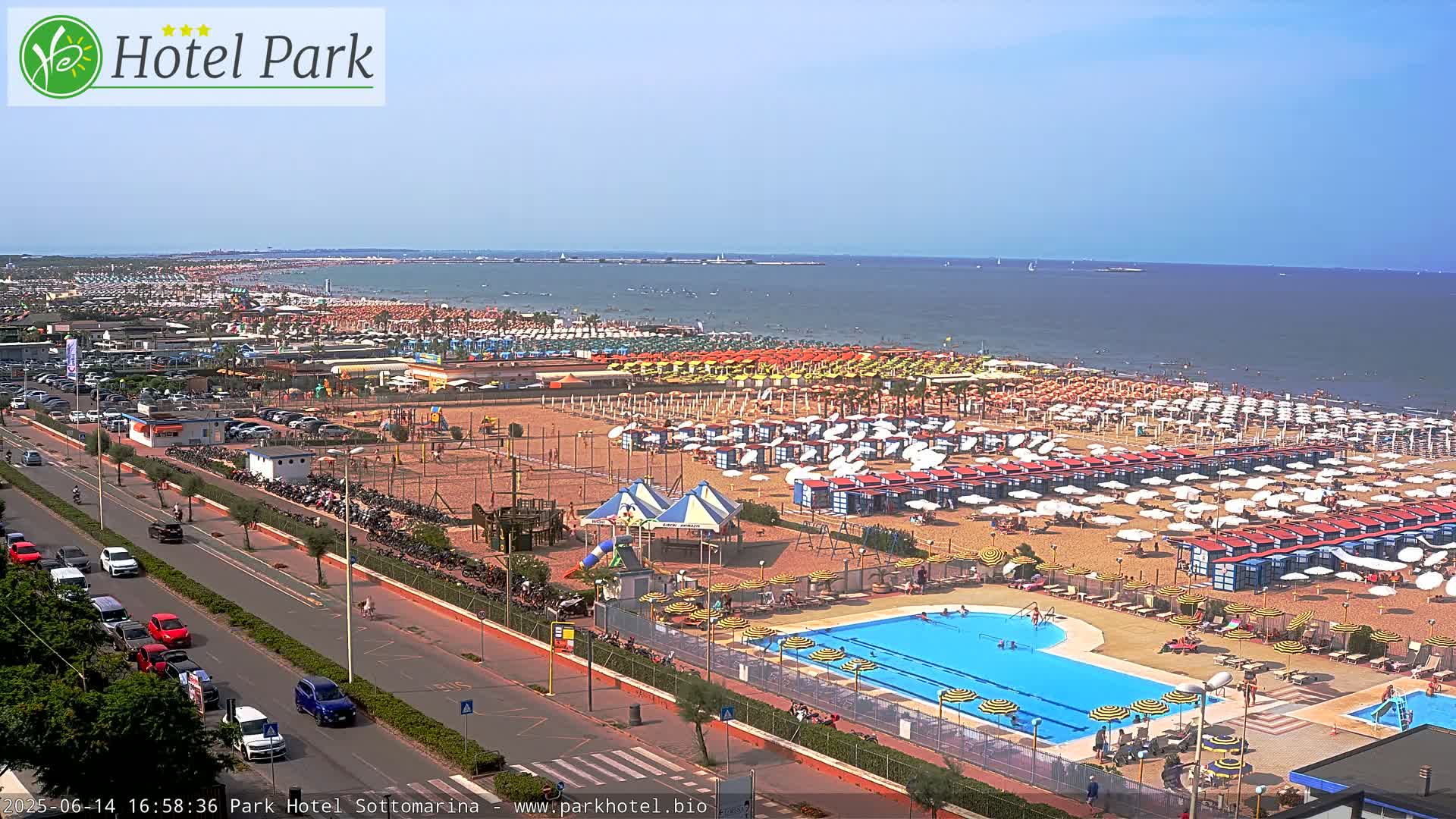 A sunny beach scene shows a crowded sandy shore with numerous umbrellas and beach chairs, a swimming pool, and a roadway with cars alongside it, all overlooking a calm ocean.