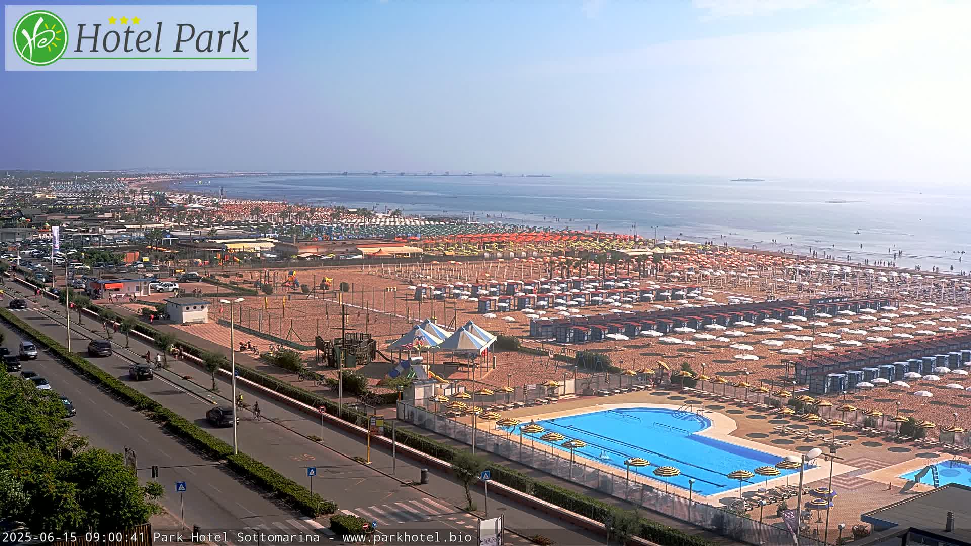 An aerial view shows a beach with numerous umbrellas and beachgoers, a swimming pool, and a roadway lined with cars under a clear, sunny sky.