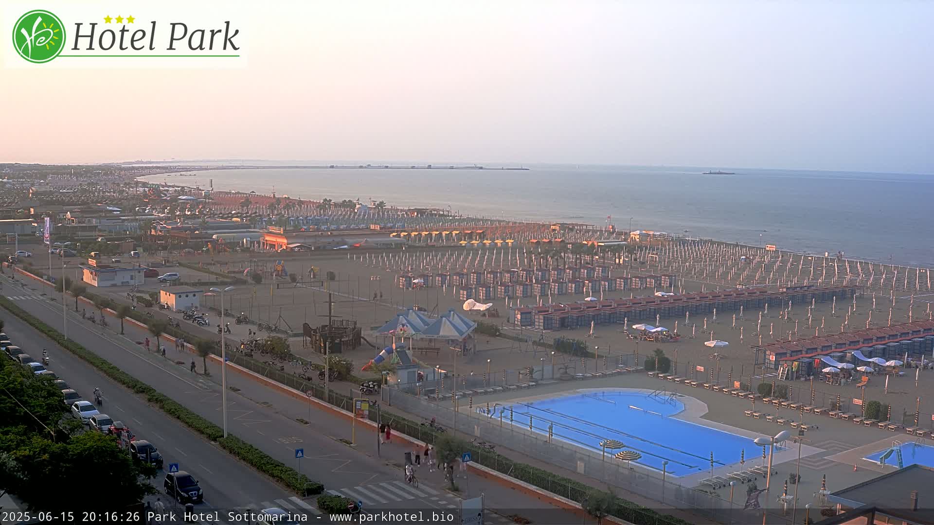An aerial view of a beach with numerous beach umbrellas and a swimming pool, overlooking a calm sea under a clear, dusky sky.