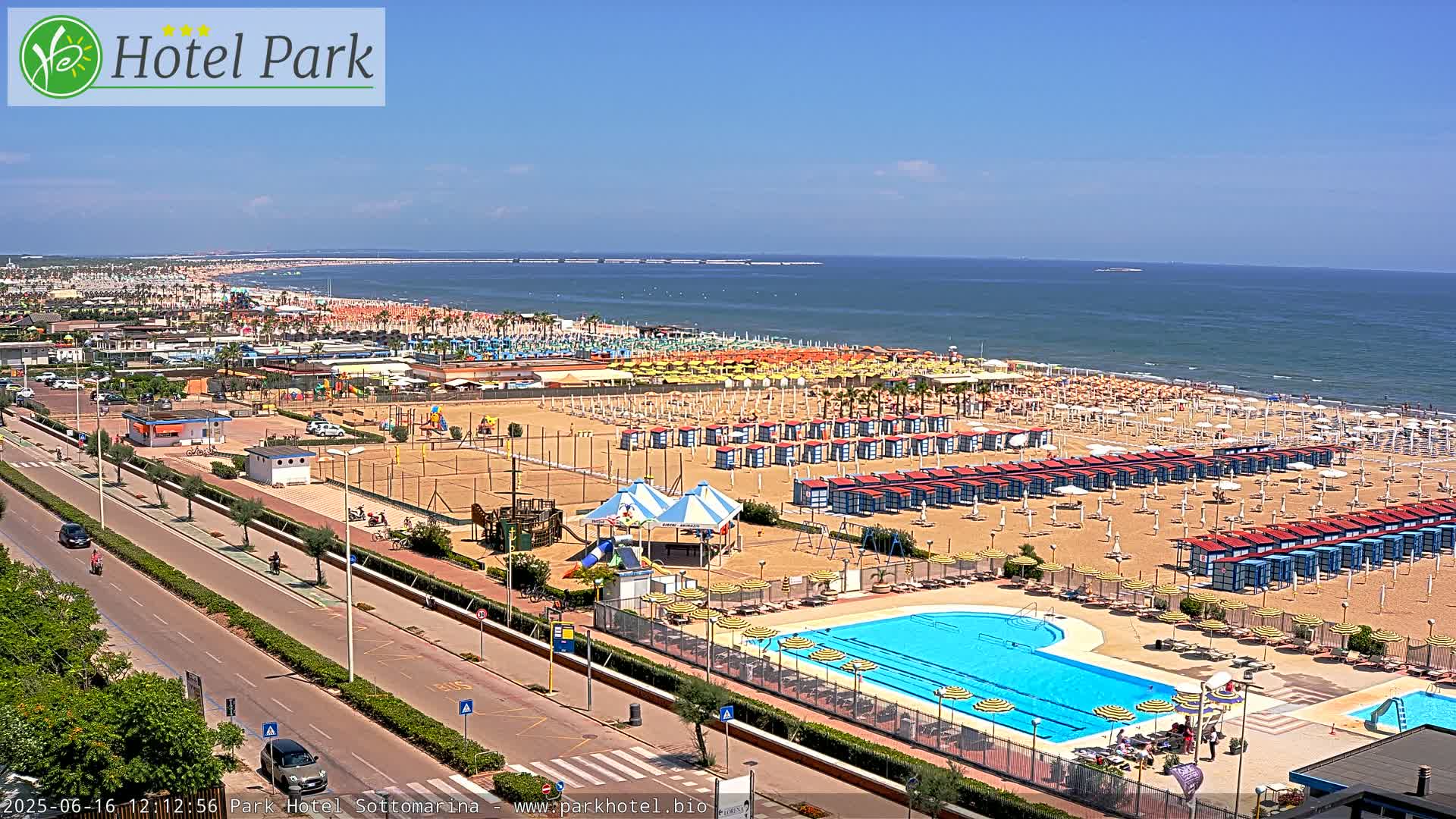 A sunny aerial view shows a beach with numerous beach umbrellas and cabanas, a swimming pool, playground equipment, and a road next to it.