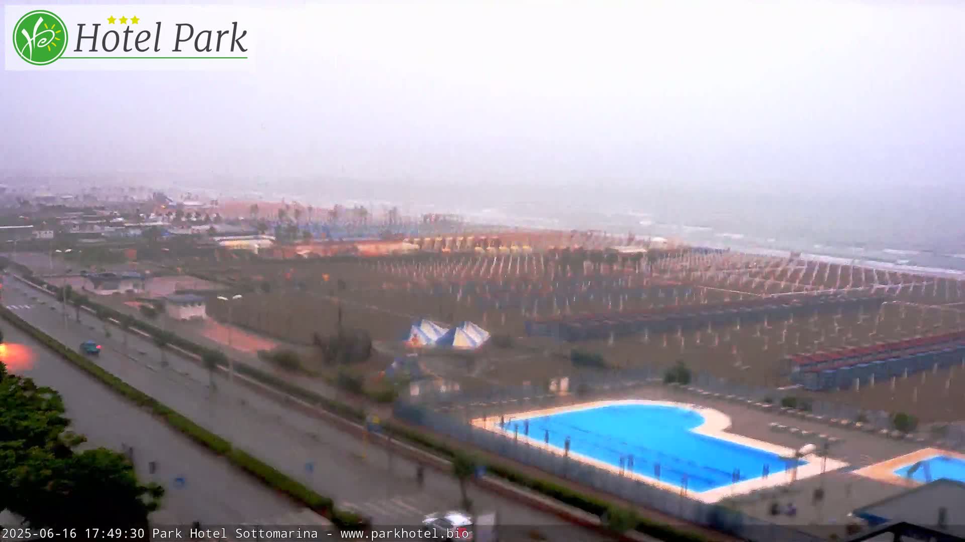 A rain-swept beach scene shows a sandy shore lined with closed beach umbrellas, a swimming pool, and a road with sparse traffic.