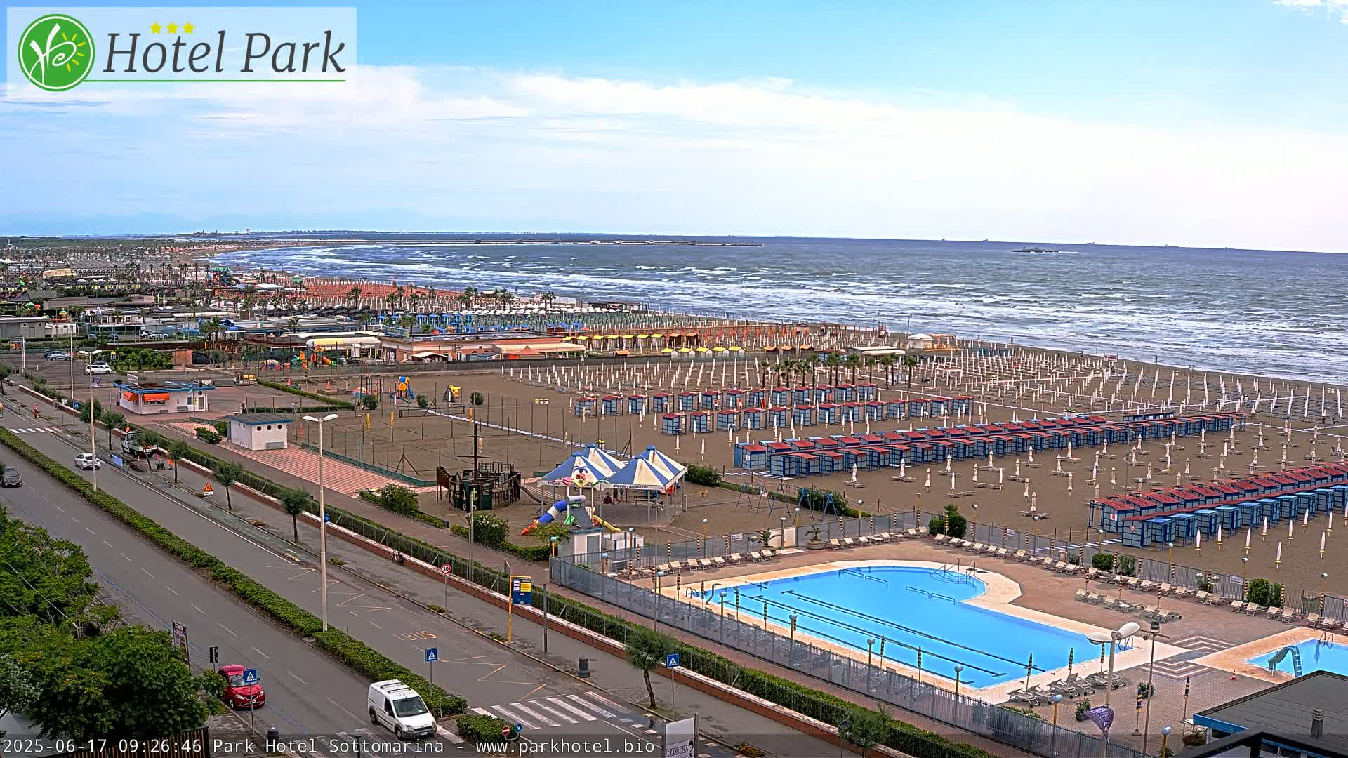 A beach with numerous beach umbrellas and a swimming pool is seen next to a road, all under a partly cloudy sky with a somewhat choppy sea.