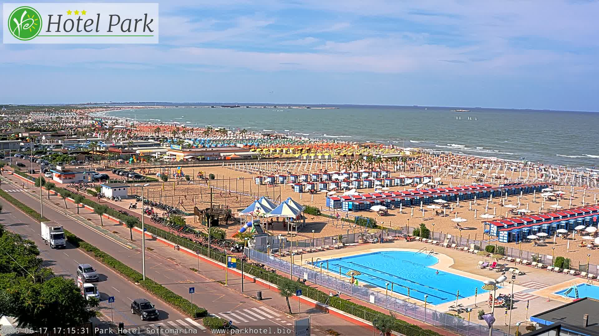 A sunny day at a beach resort shows a long sandy beach lined with rows of beach umbrellas and cabanas, a swimming pool, and a roadway leading to a parking lot.