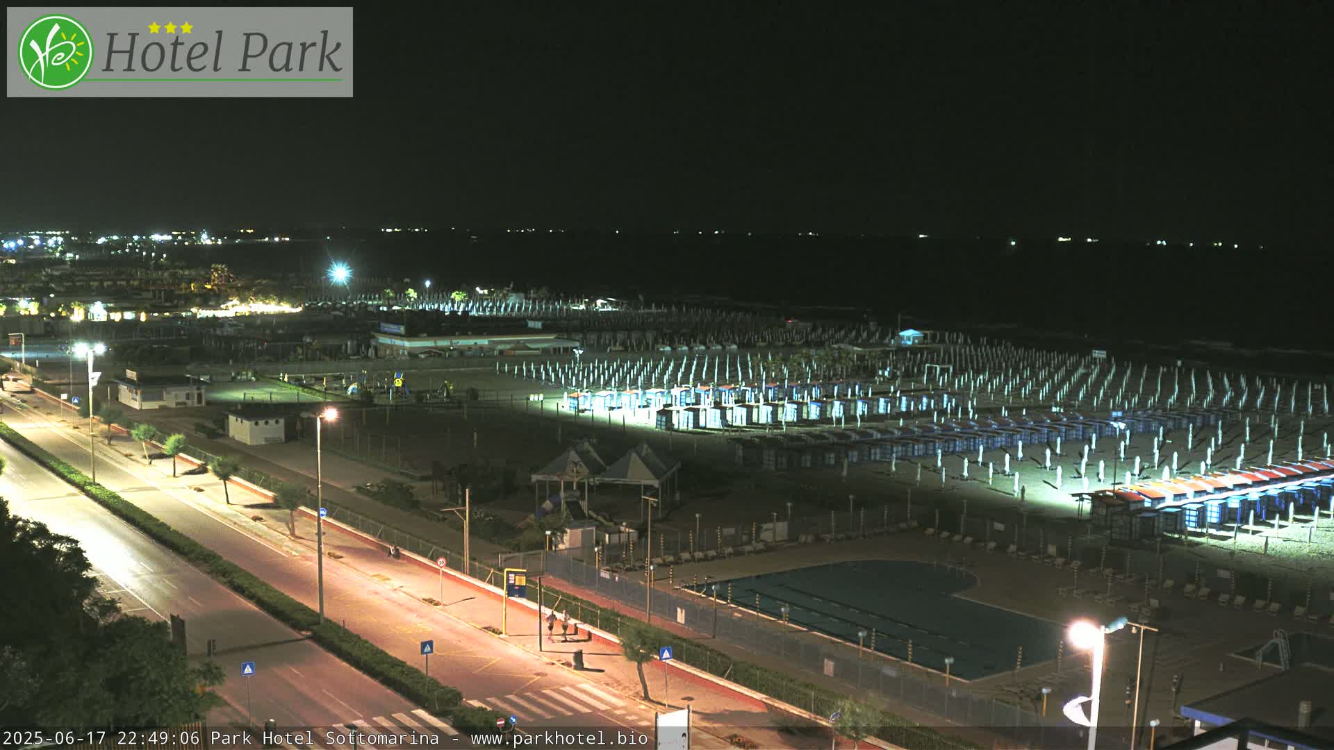 A nighttime aerial view of a beach with numerous closed beach umbrellas and a swimming pool, all illuminated under a dark sky.