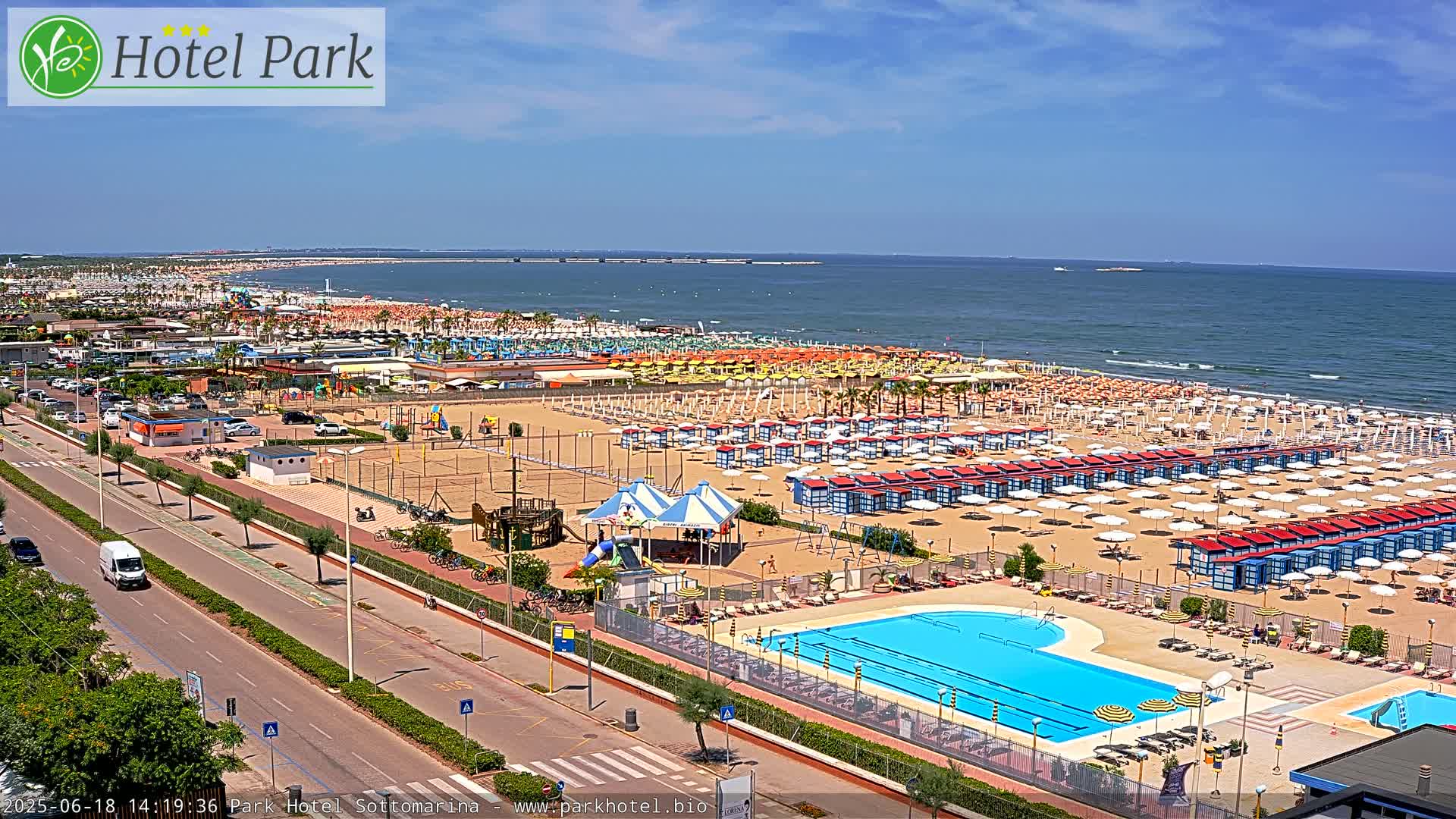 A sandy beach with numerous umbrellas and beach cabanas, a swimming pool, playground equipment, and a roadway, all under a clear, sunny sky.