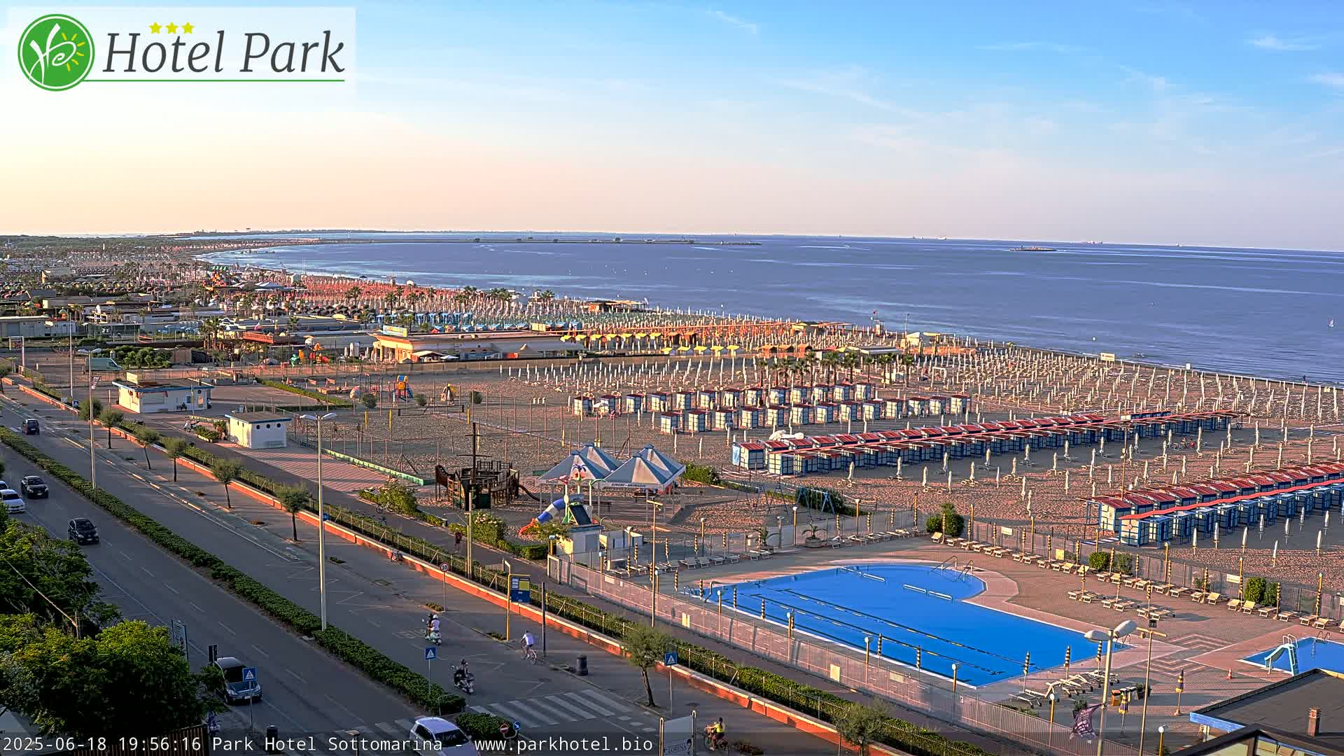 An aerial view shows a beach with numerous beach umbrellas and cabanas, a swimming pool, a playground, and a road leading to the ocean under a clear, bright sky.