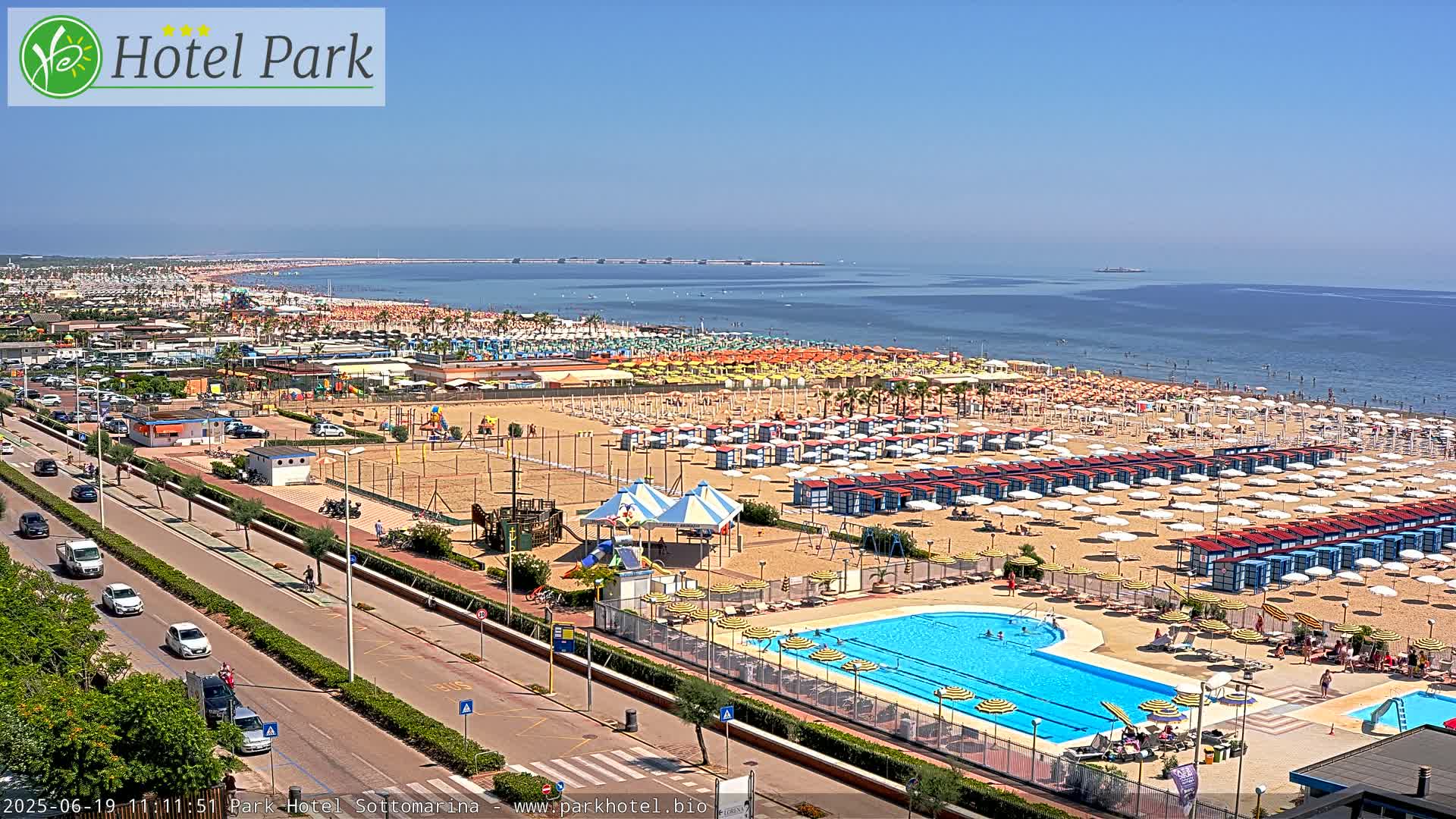 A sunny day at a beach resort shows a long sandy beach with numerous umbrellas and beach cabanas, a swimming pool, and a road with cars driving alongside it.