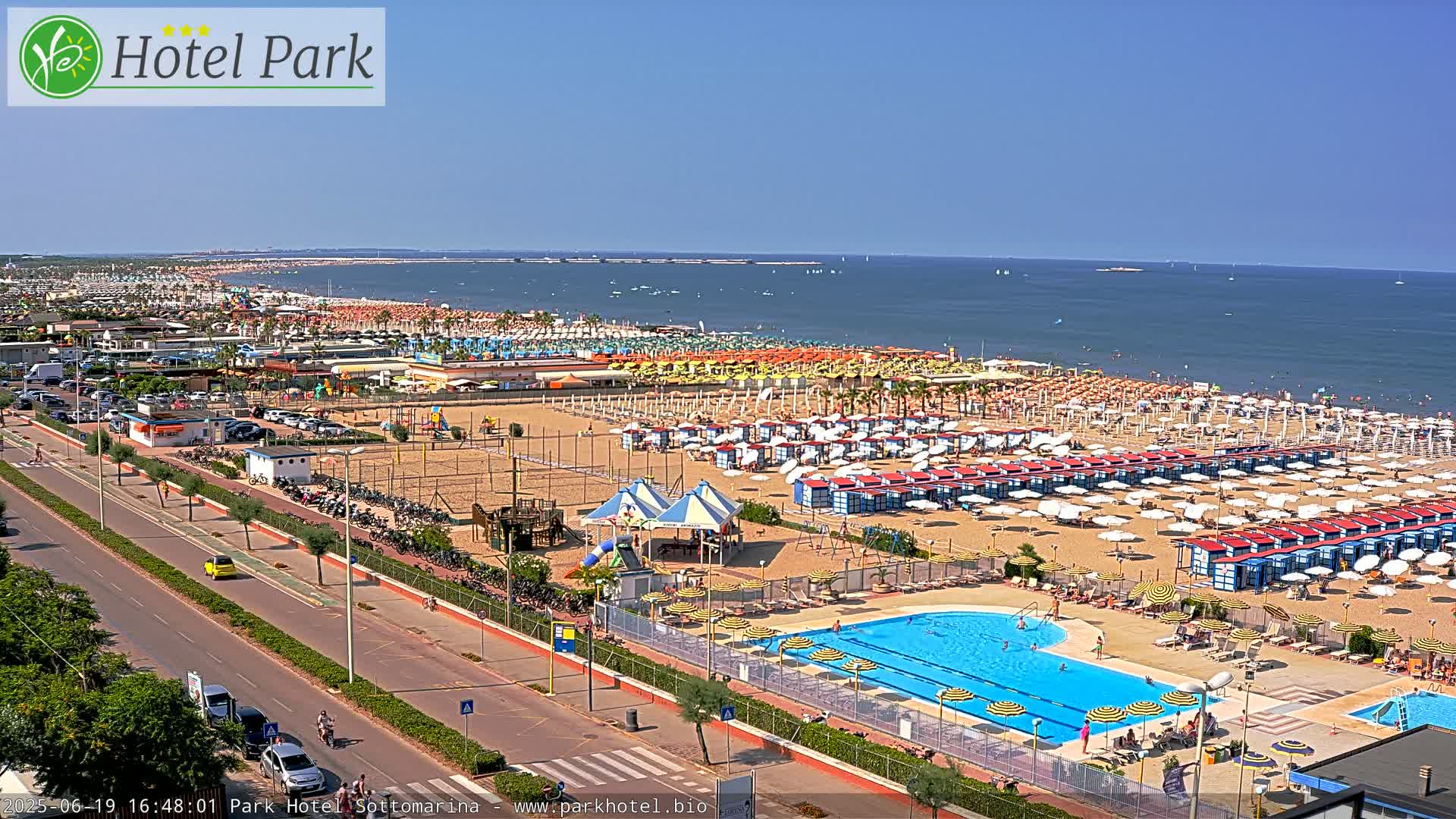 A sunny beach scene shows a sandy shore lined with rows of beach umbrellas and cabanas, a swimming pool, and a road with cars.