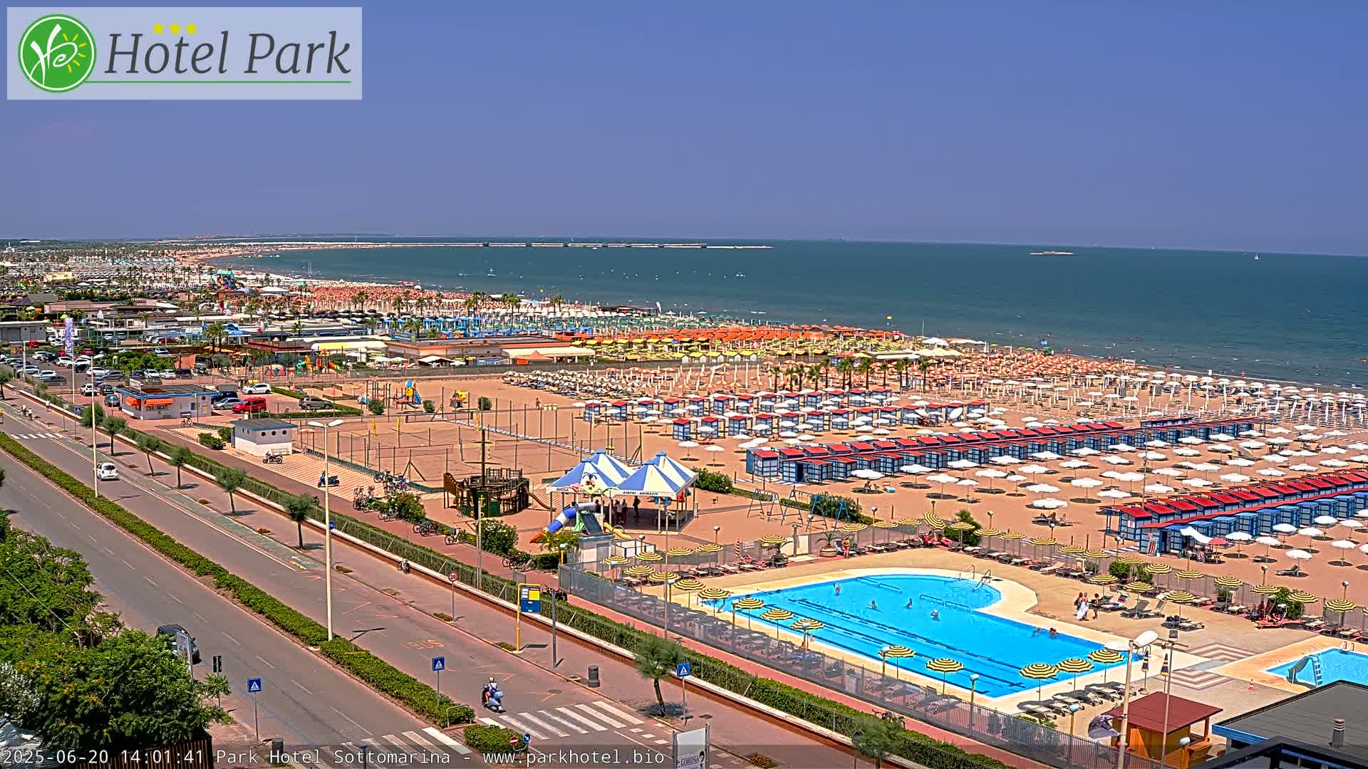 A sunny beach scene shows a long sandy shore lined with numerous beach umbrellas and a swimming pool, with a road and buildings visible beyond.