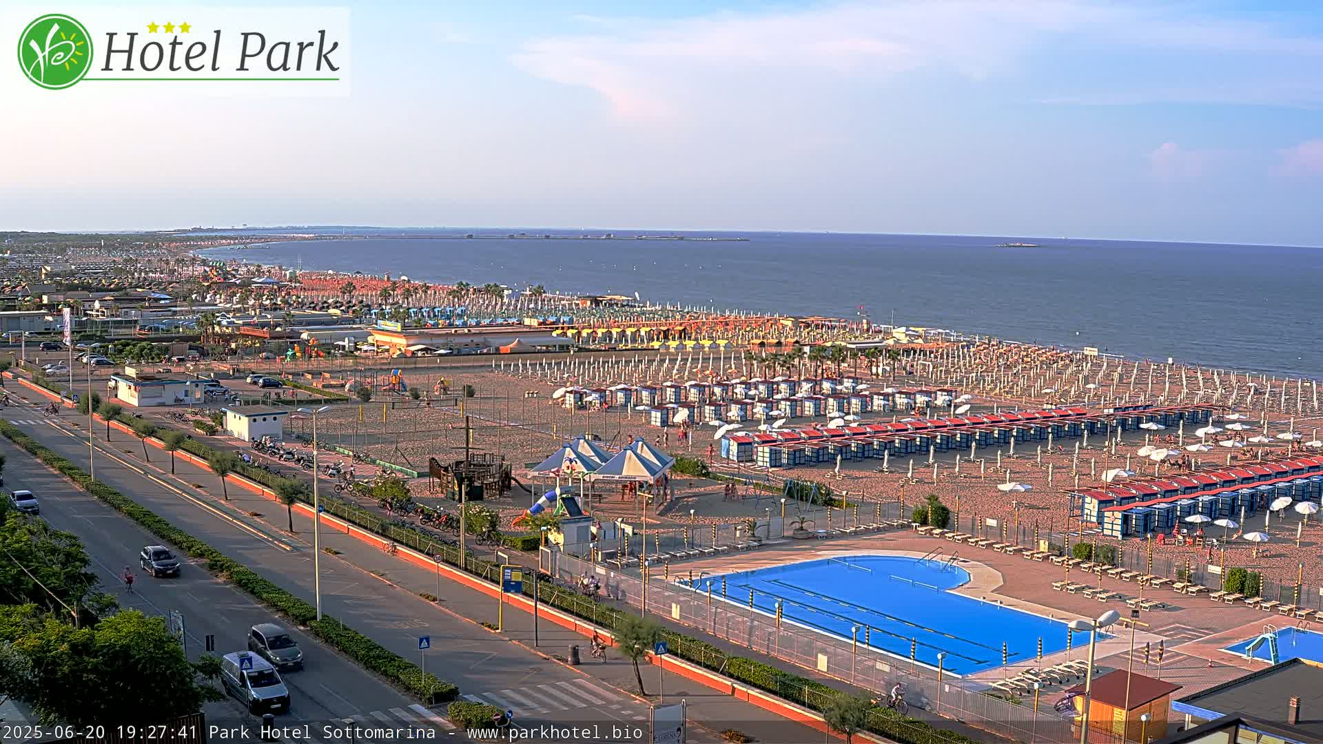 A sandy beach with numerous beach umbrellas and changing rooms, a swimming pool, playground, and a road beside it, all under a clear, sunny sky.