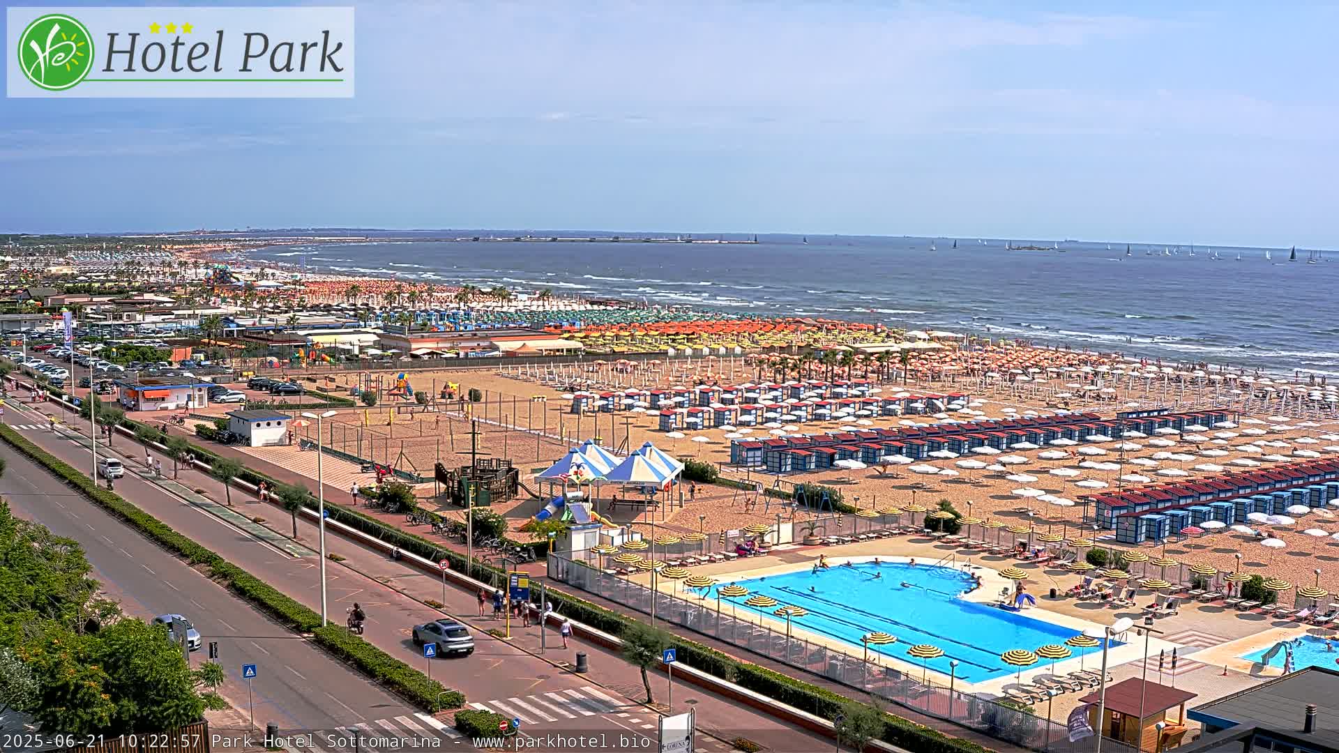 A sunny day at a beach resort shows a wide sandy beach crowded with umbrellas and beachgoers, a swimming pool, and a road running parallel to the shore.